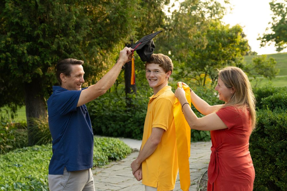 Parents placing graduation cap on smiling teen son outdoors in green park setting.