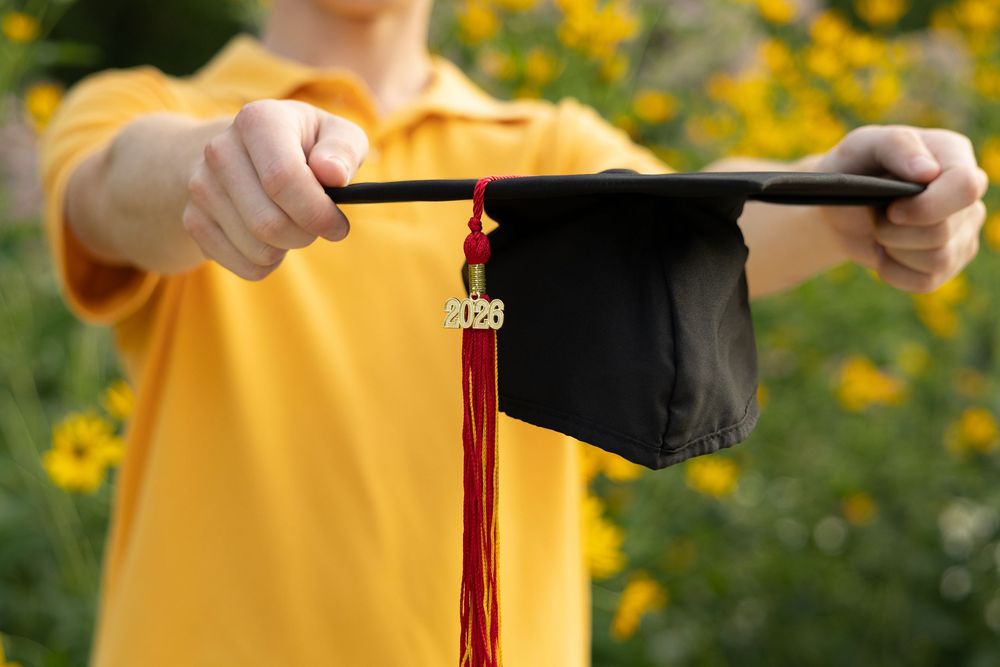 Person in yellow shirt holding a black 2026 graduation cap with red tassel outdoors with yellow flowers in background.