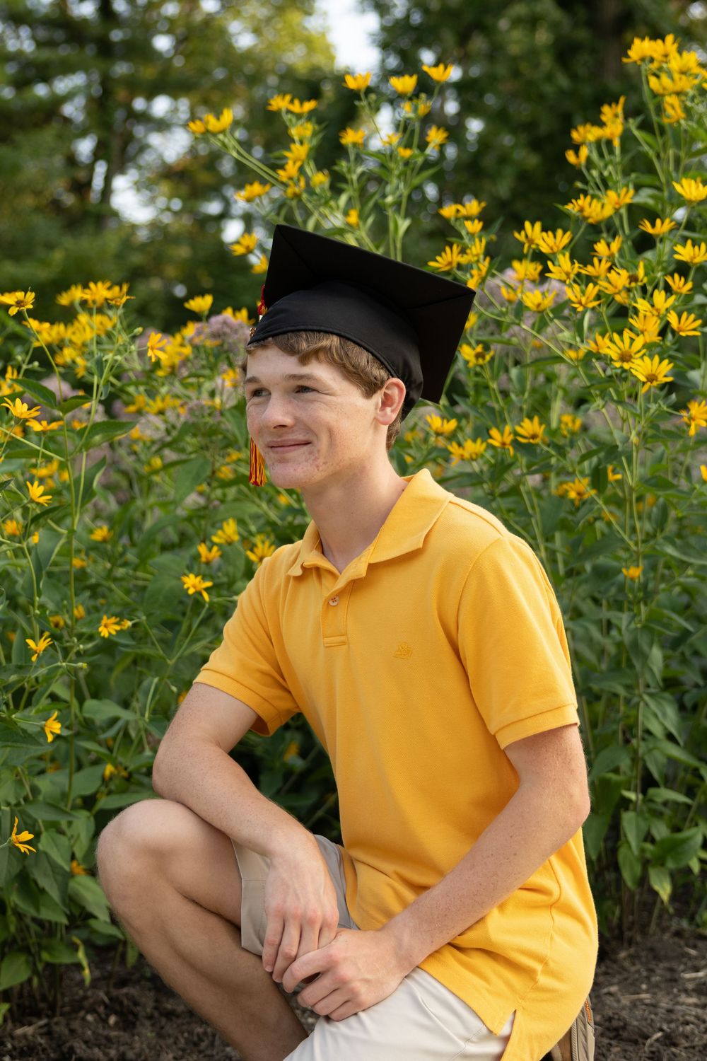 Young man in yellow polo shirt and graduation cap smiles outdoors among bright yellow wildflowers.