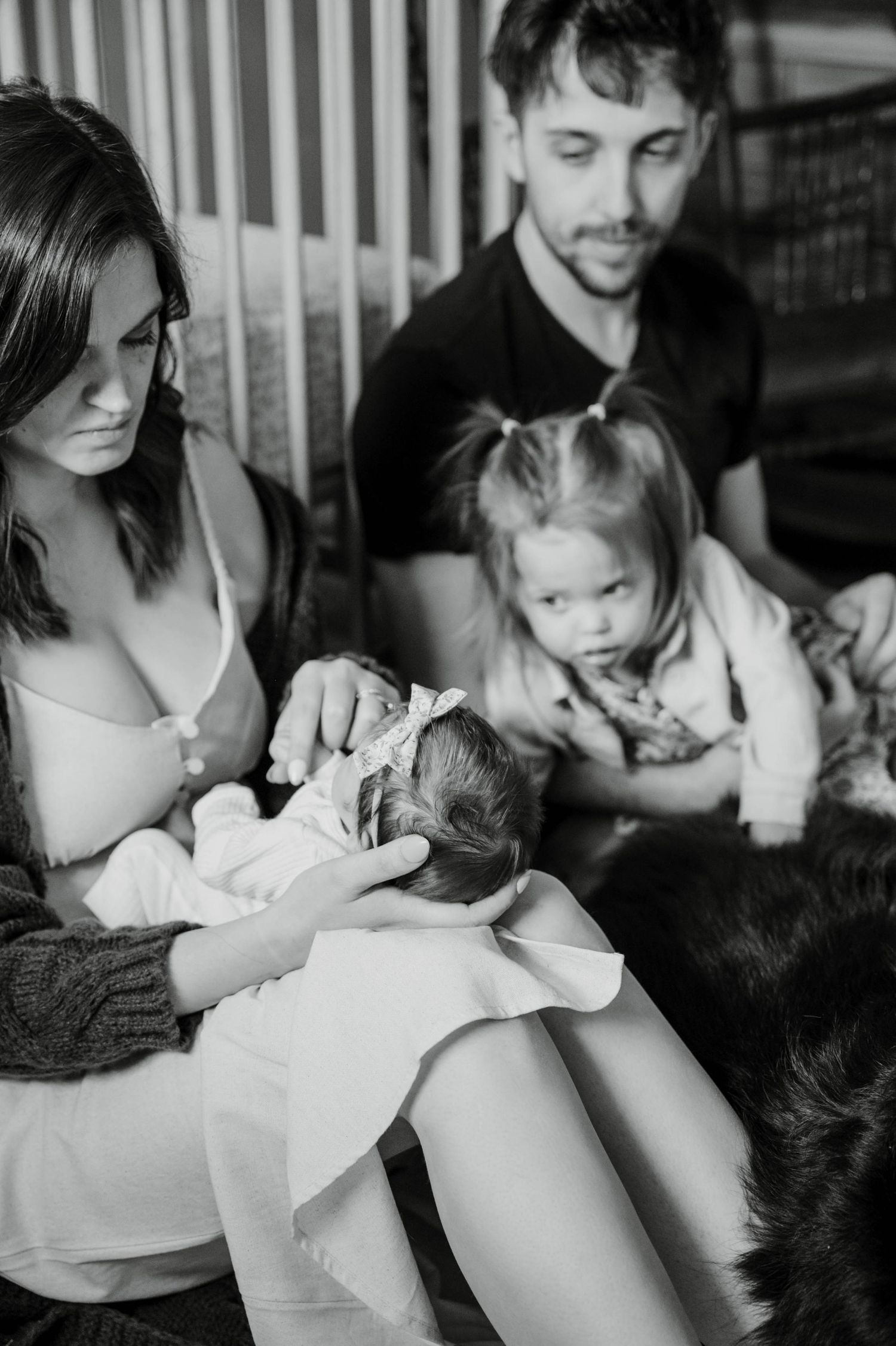A black and white portrait of a family sitting together with a young child and an infant in a casual indoor setting.