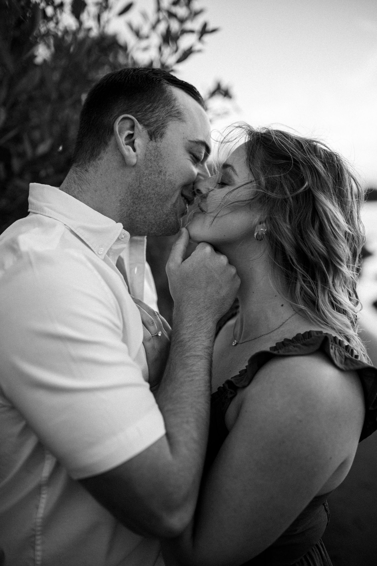 A romantic black and white photograph captures an intimate kiss between a couple outdoors.