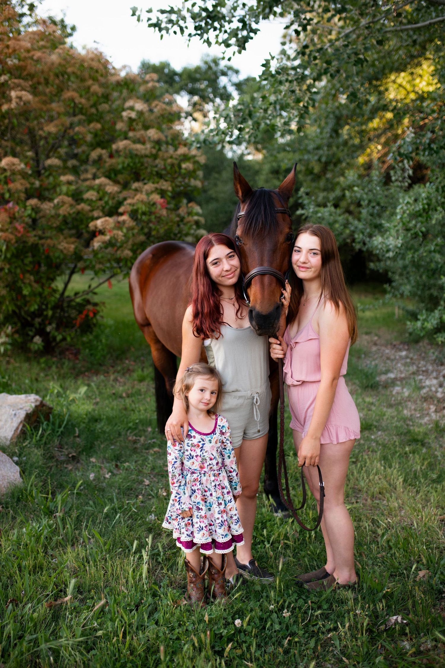Three smiling girls and a horse standing together outdoors in a lush green garden field in Nevada City.