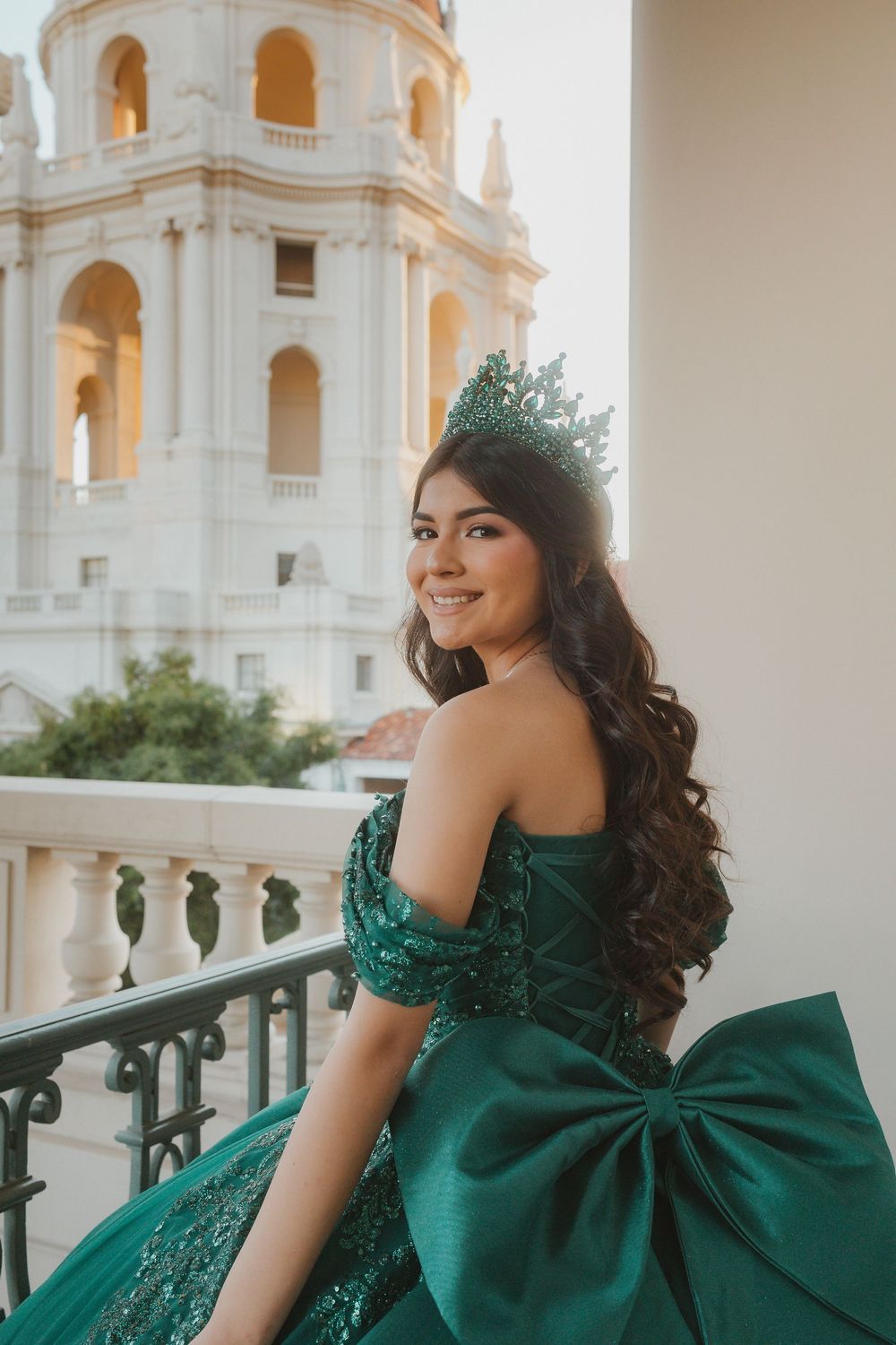 An emerald green strapless ballgown with a large bow detail shown against a white architectural backdrop.