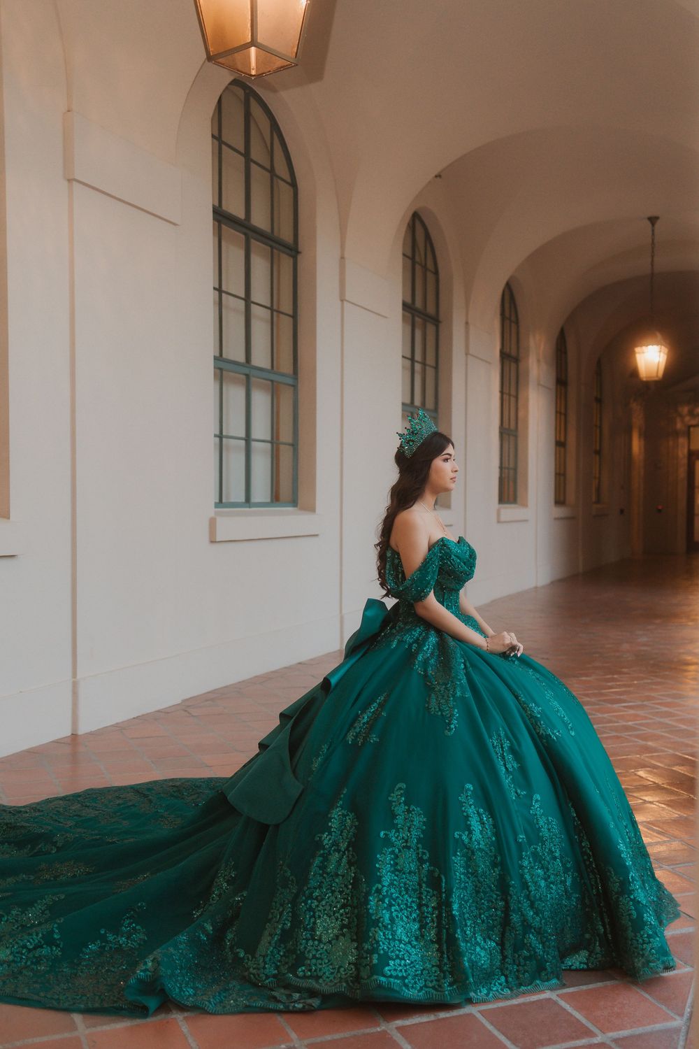 A stunning emerald green ball gown with intricate lace details sweeps across a tiled corridor with arched windows.