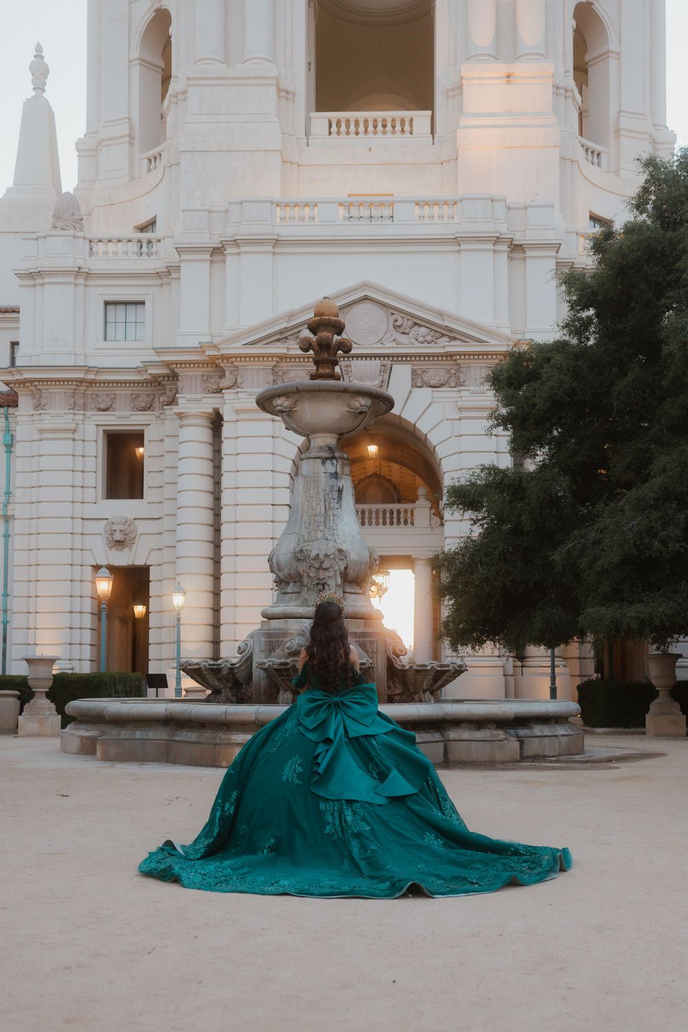 A grand teal ballgown dress spreads across marble steps in front of an ornate white classical building with fountain.