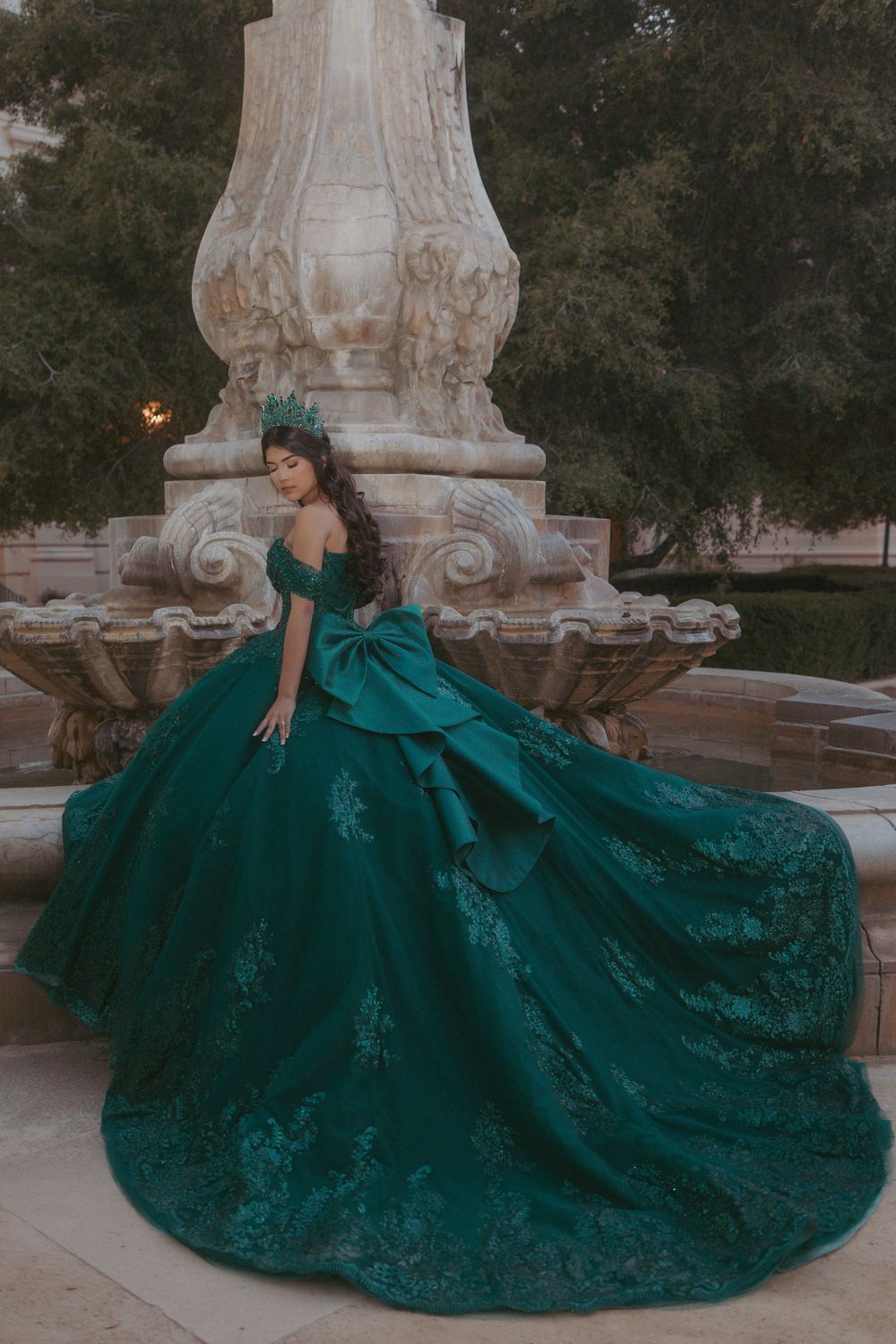 Stunning emerald green ball gown with intricate lace details photographed against an ornate stone fountain in an outdoor setting.
