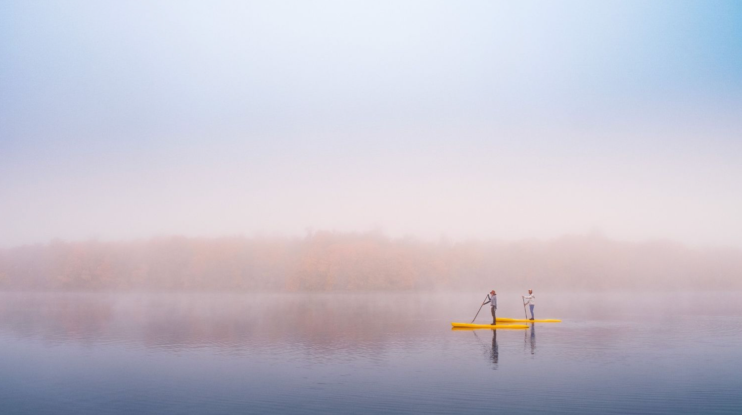 A pair of Stand-up paddleboarders glide across misty lake waters during a tranquil sunrise with hazy mountains in background.