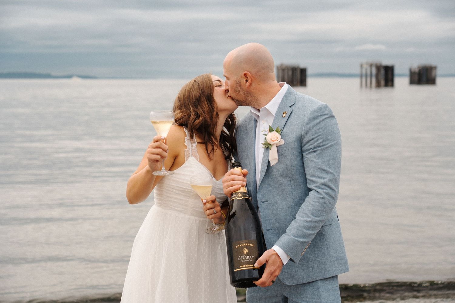 Newlywed couple shares a romantic moment with champagne on a cloudy beach with wooden pier in background.