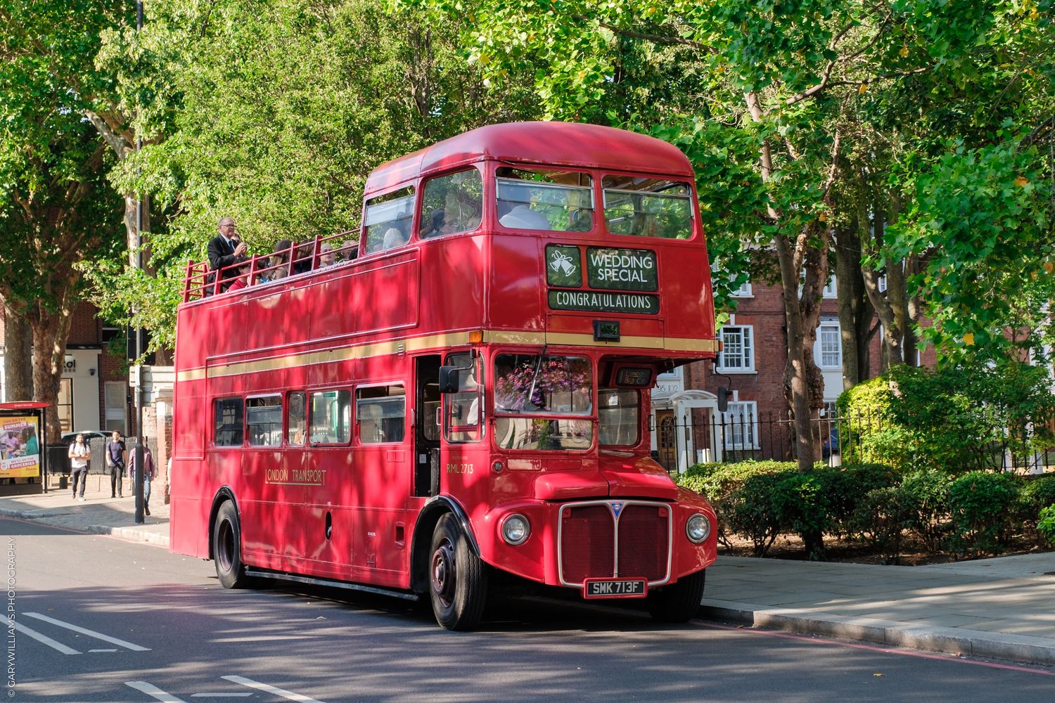 A classic red double decker Routemaster bus parked along a leafy London street during a sunny summer day.