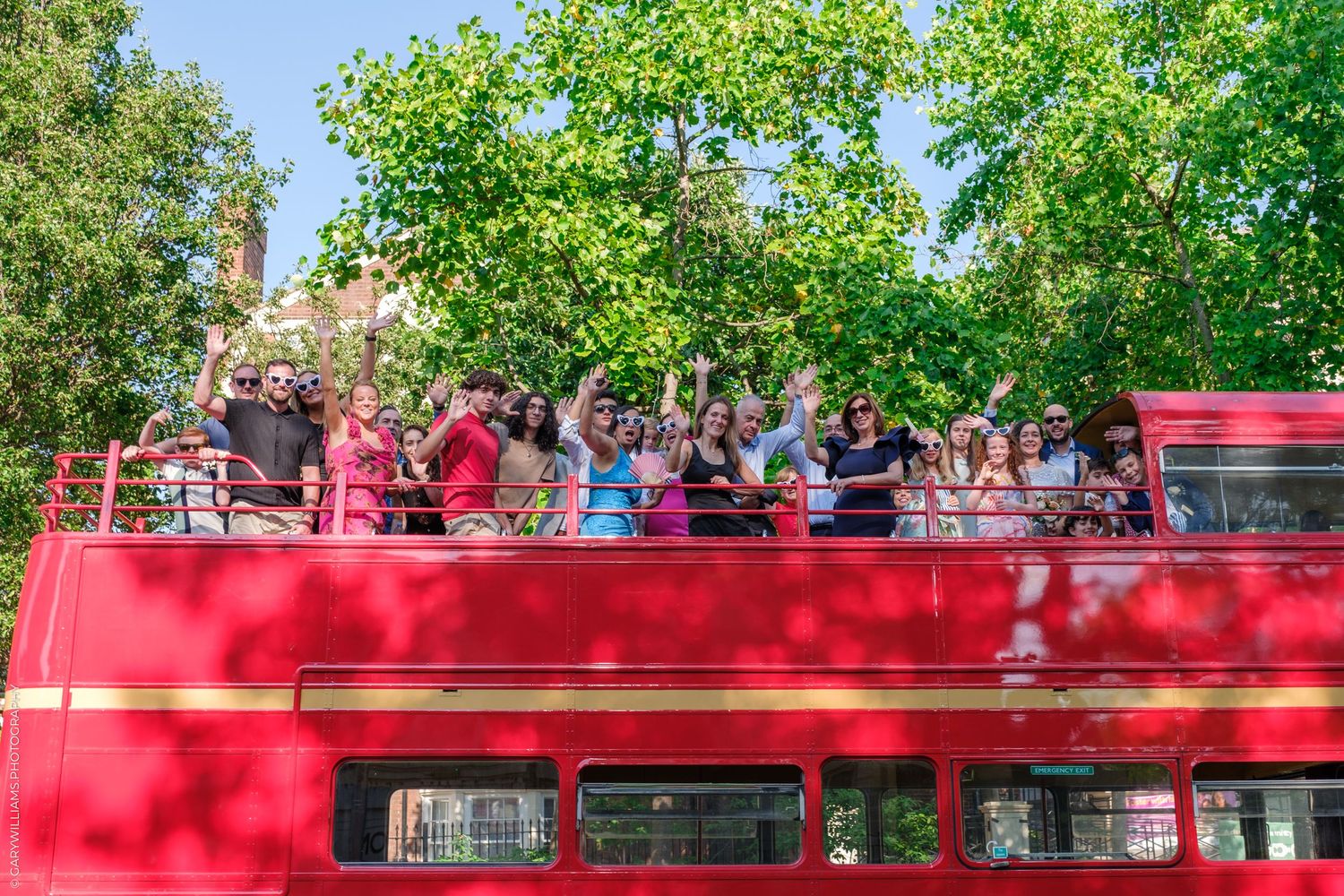 Red double decker bus parked under lush green trees on a sunny day.