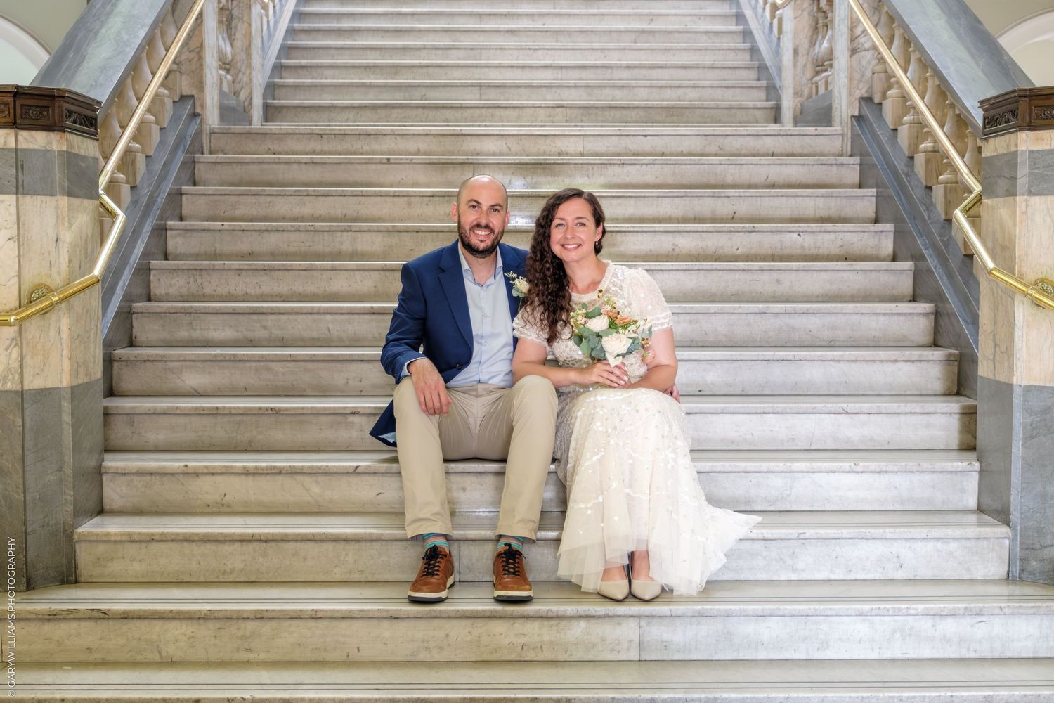 A couple sits together on white marble stairs, the bride wearing a lace dress and holding a bouquet of flowers.