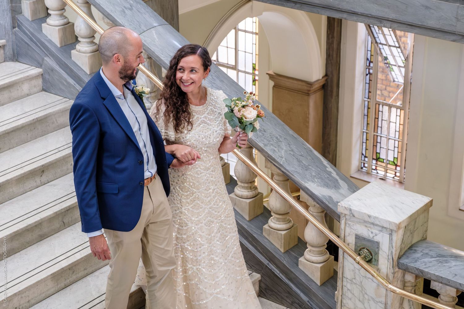 A couple shares a tender moment on an elegant stone staircase with ornate bannisters in a historical building.