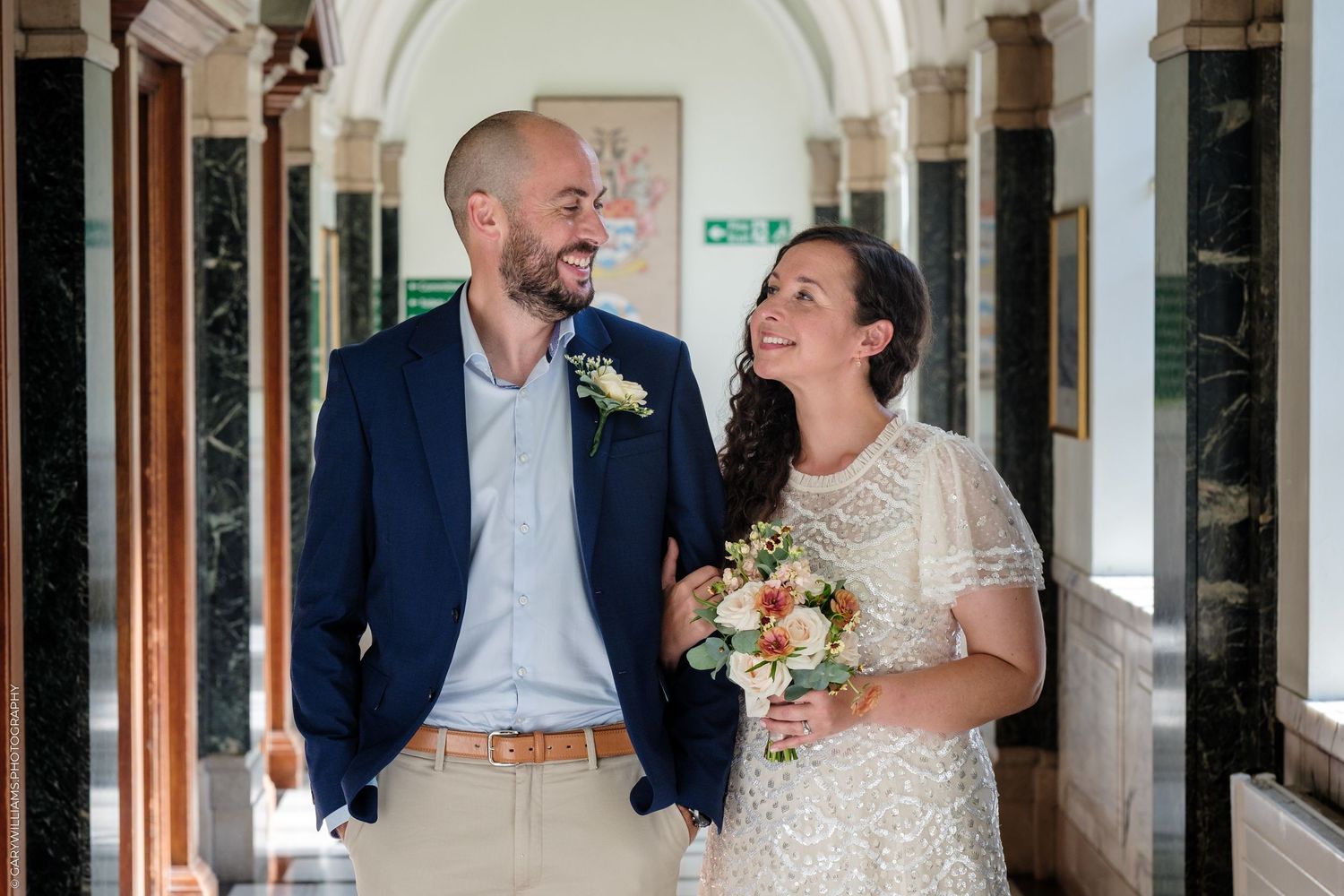 A bride and groom share a joyful laugh together while standing in an elegant arched hallway.