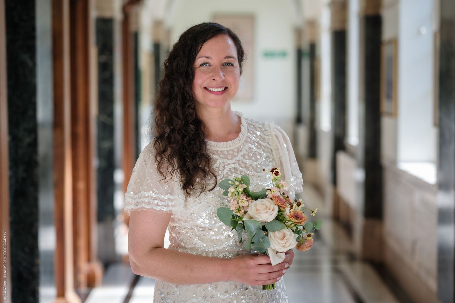 Bride in a lace wedding dress stands in a sunny hallway holding a peach and white bouquet of roses.