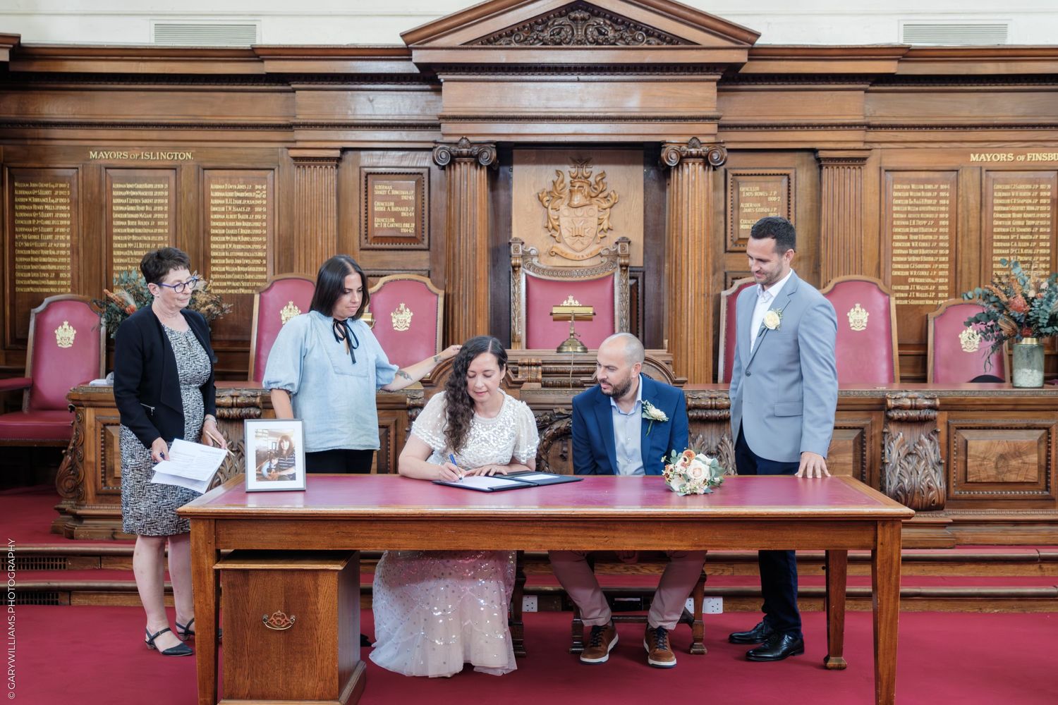Couple signing marriage documents in an ornate wood-paneled council chamber with red carpet and witnesses present.