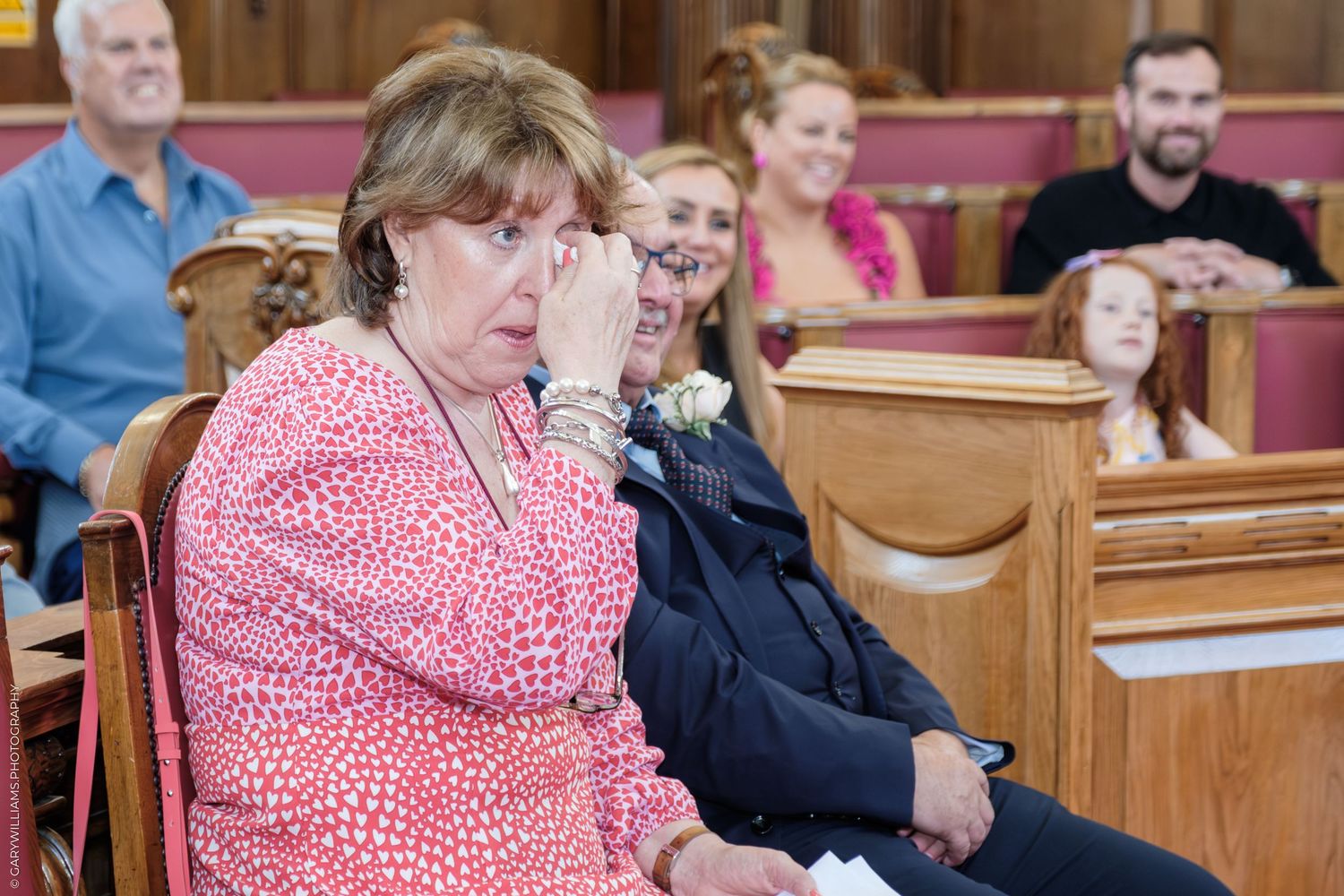 People sit in formal wooden chairs and pews in what appears to be a church or meeting hall interior.
