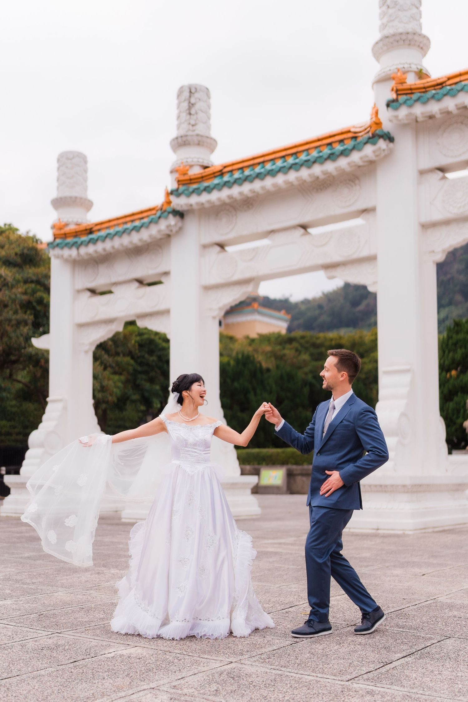 A couple dances together in formal wedding attire in front of ornate white stone pillars with traditional Asian architecture.