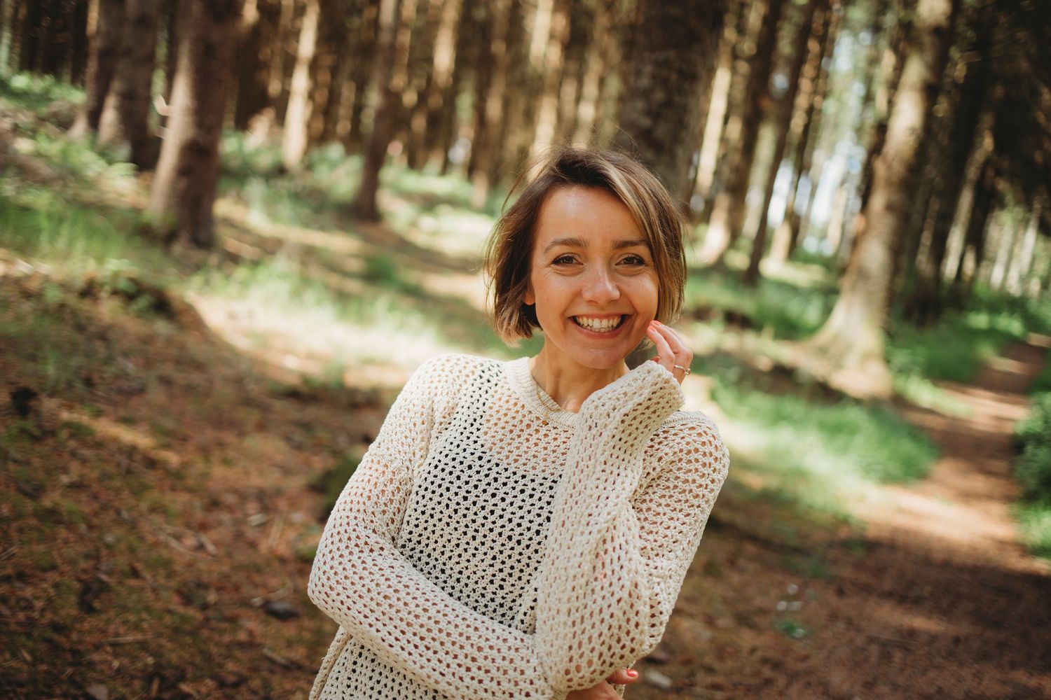 Someone wearing a cozy white knit sweater smiles while standing in a sunlit forest path.