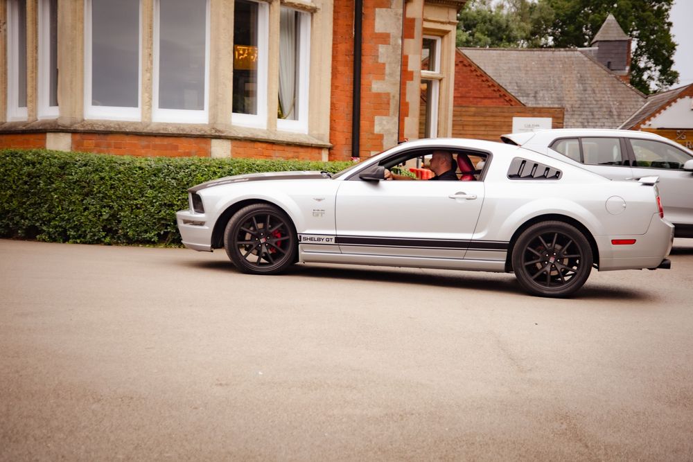 A silver Ford Mustang GT with custom wheels parked on a residential street next to brick buildings.