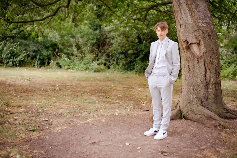 A person wearing a white suit and sneakers stands next to a large tree trunk in a natural outdoor setting.
