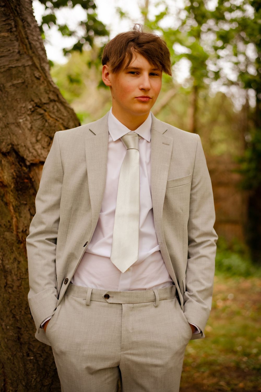 Portrait of someone in a light gray suit and white tie standing near a tree trunk in an outdoor setting.