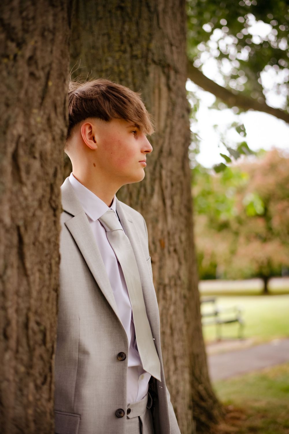 A person in a light grey suit jacket poses thoughtfully against a tree trunk in an outdoor park setting.