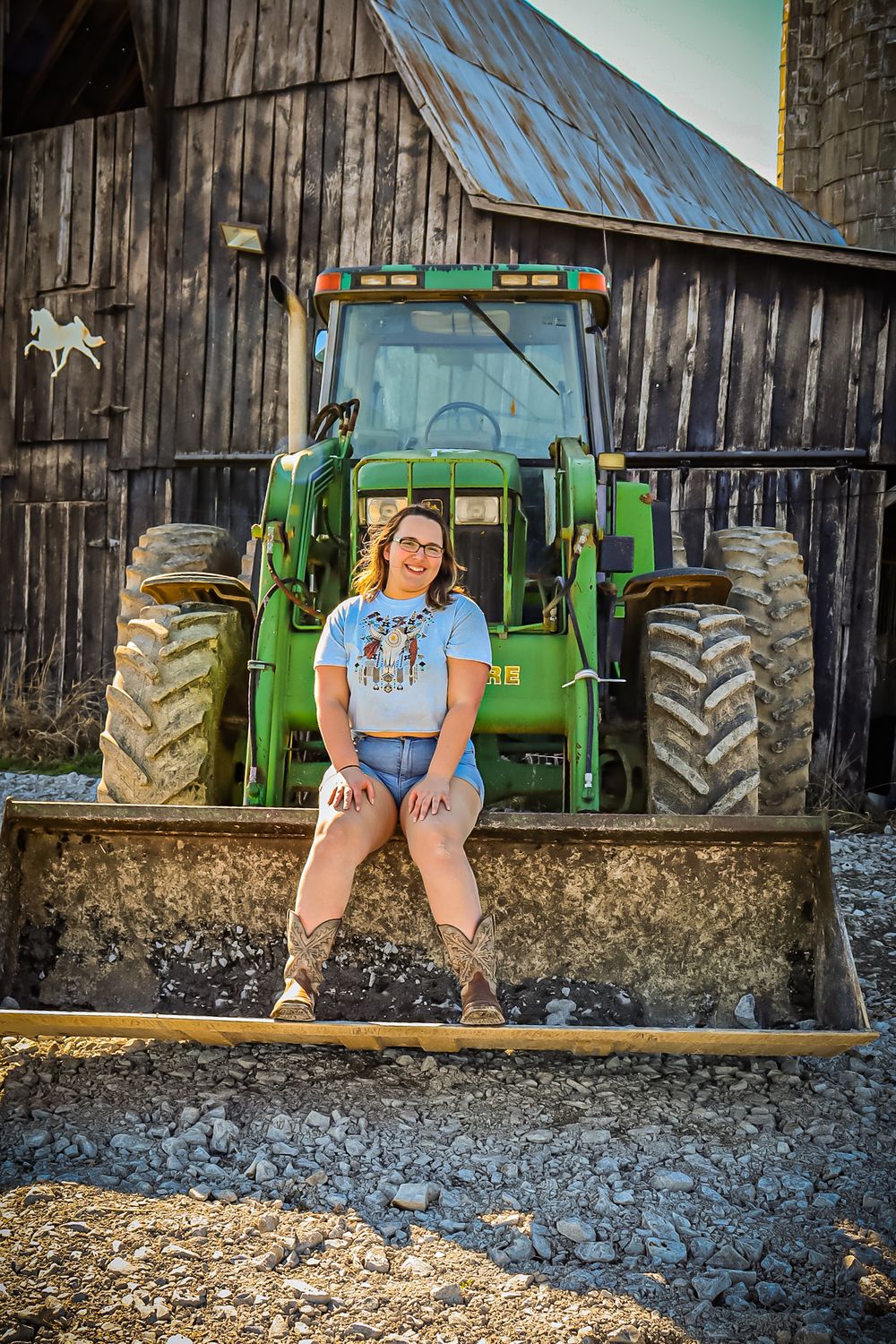 A green John Deere tractor parked in front of a rustic wooden barn with someone sitting in its front loader bucket.