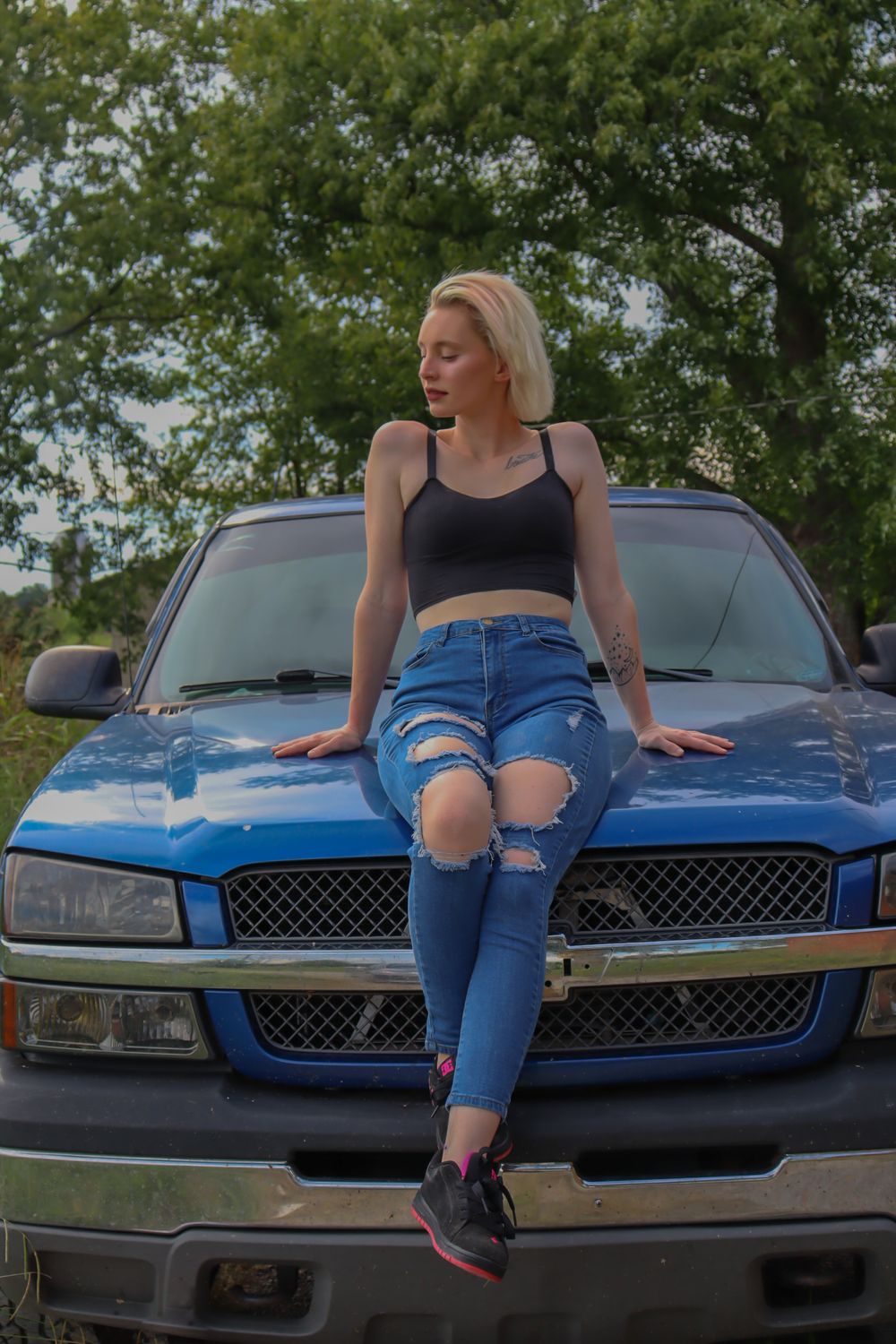 A person in ripped jeans and crop top sits on the hood of a blue Chevrolet truck against a leafy background.