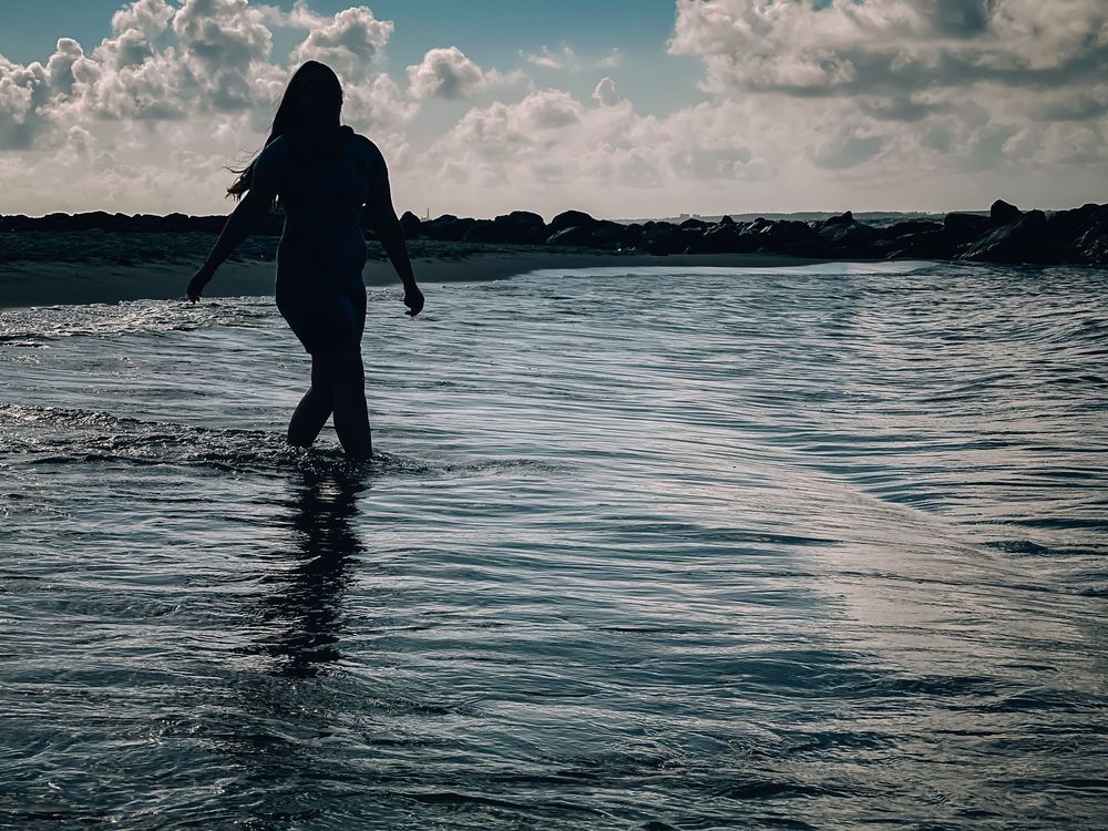 A silhouetted figure wades through shallow ocean water at sunset with cloudy skies above.