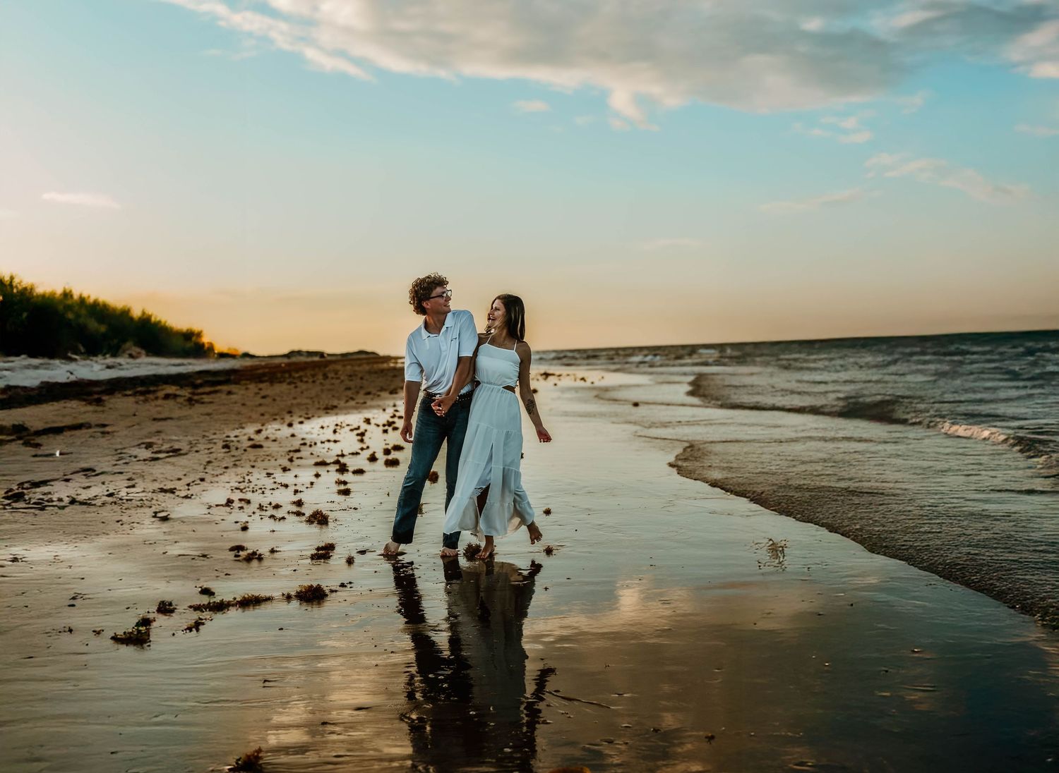 Two figures walk together on a beach at sunset with waves washing over the sand.