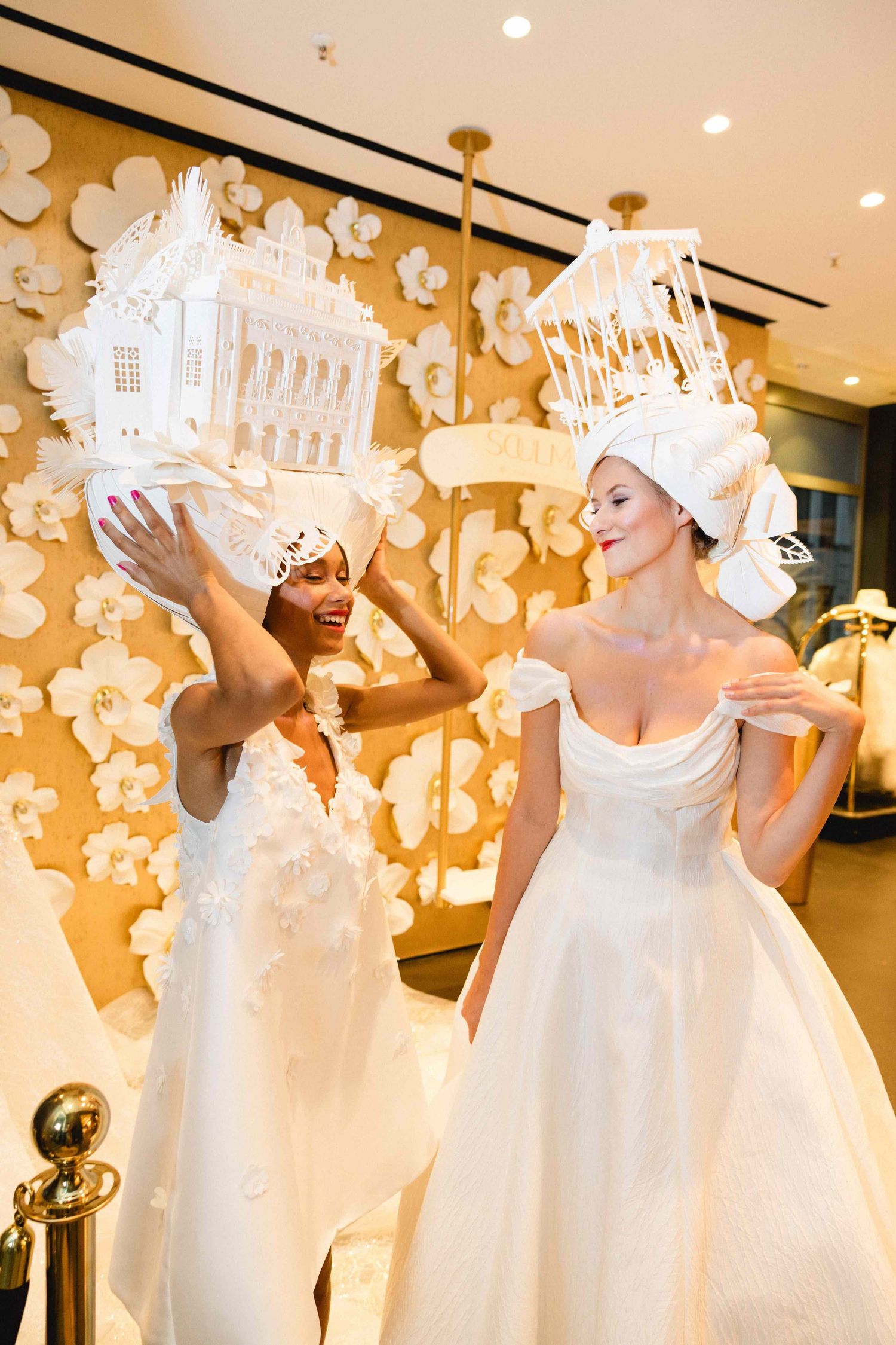Two people in elegant white wedding gowns interact playfully beneath ornate architectural headpieces in a bridal boutique.