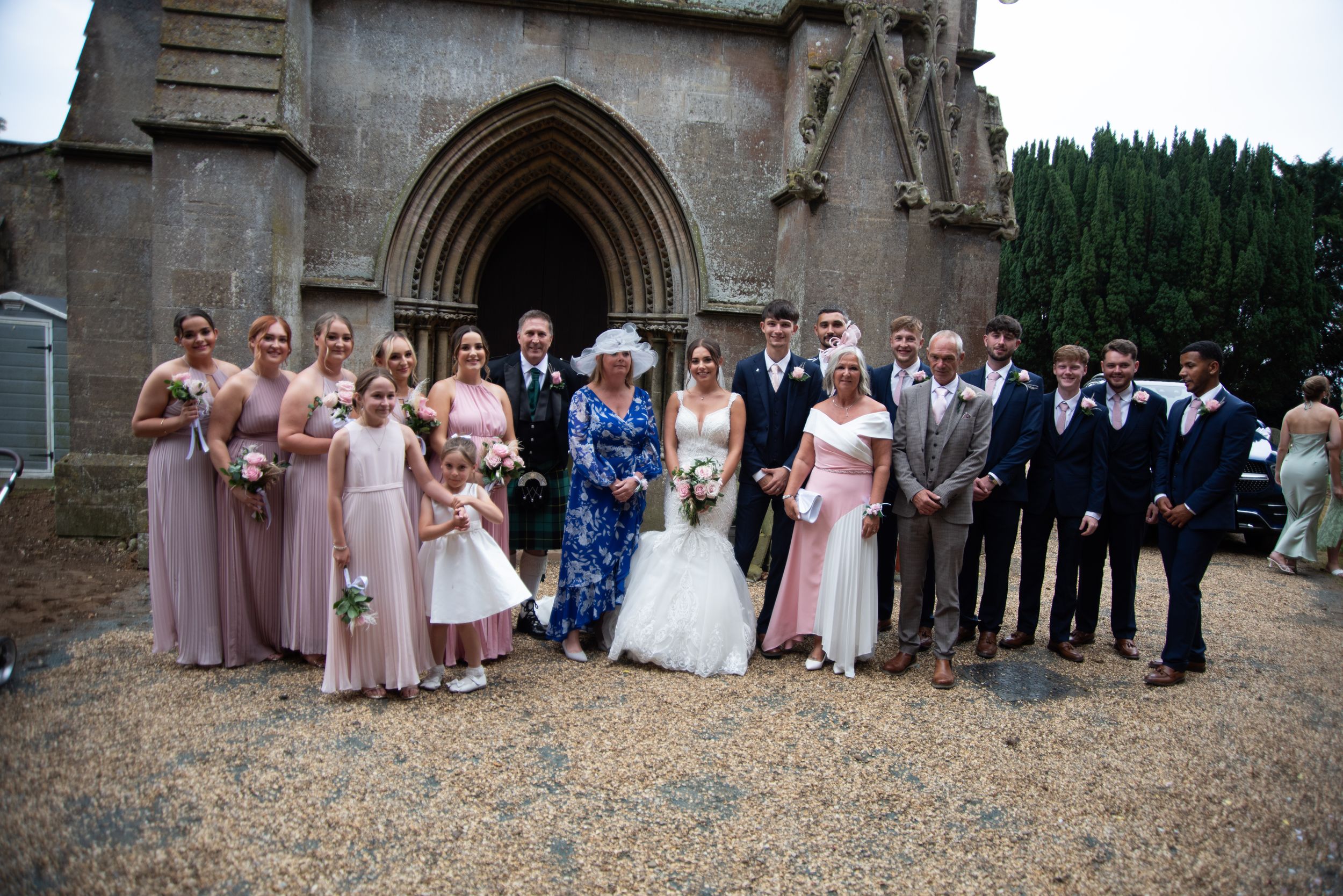 Wedding party poses together outside historic stone church with Gothic arched doorway on gravel path.
