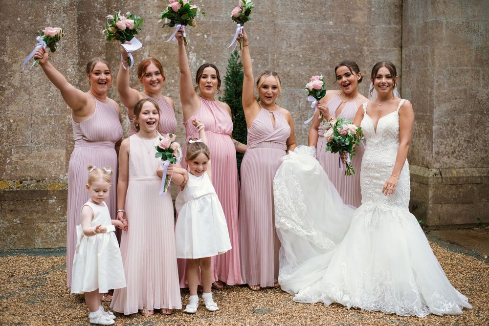 A wedding party in blush pink dresses holding bouquets poses together outdoors against a stone wall.