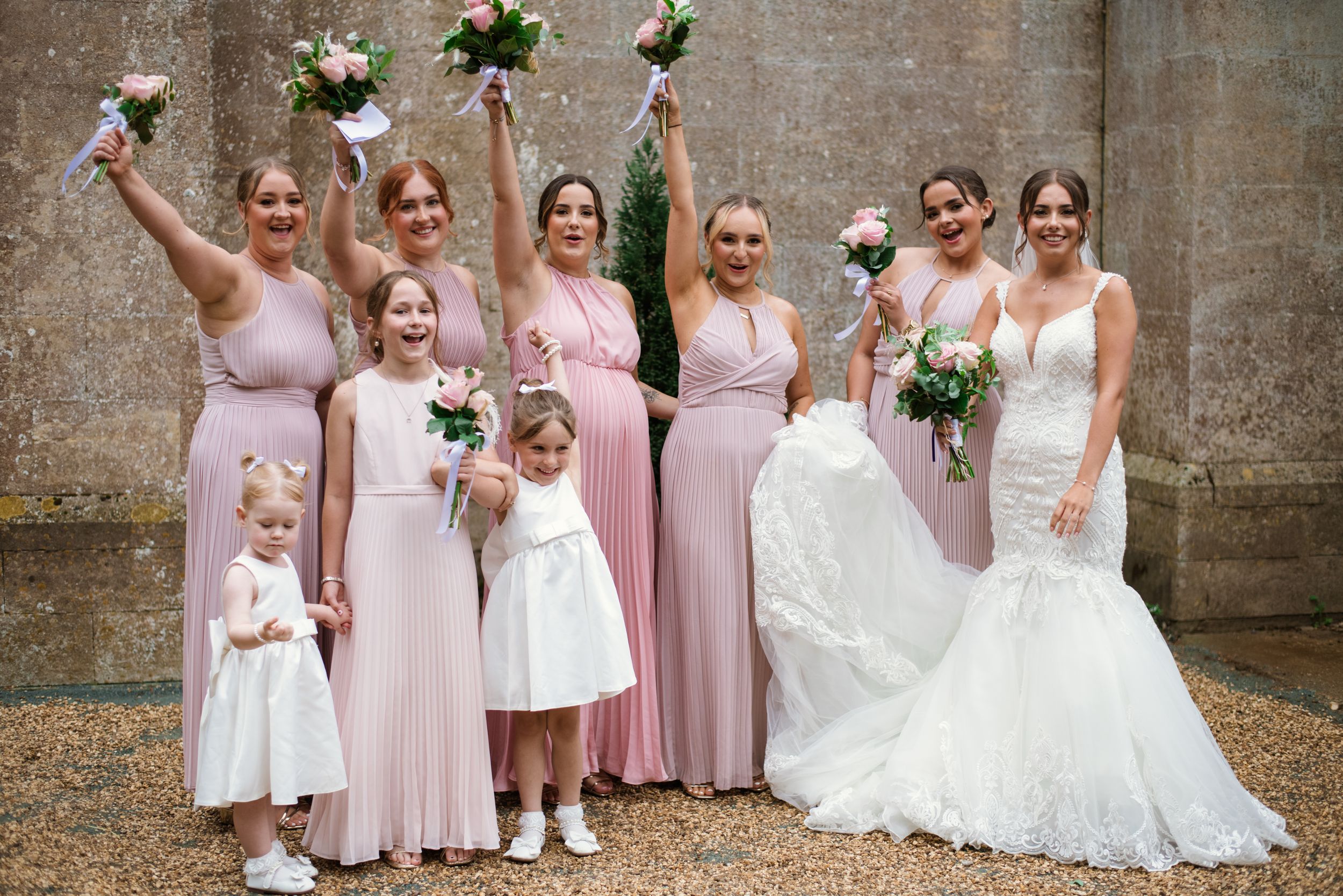 A wedding party in blush pink dresses holding bouquets poses together outdoors against a stone wall.