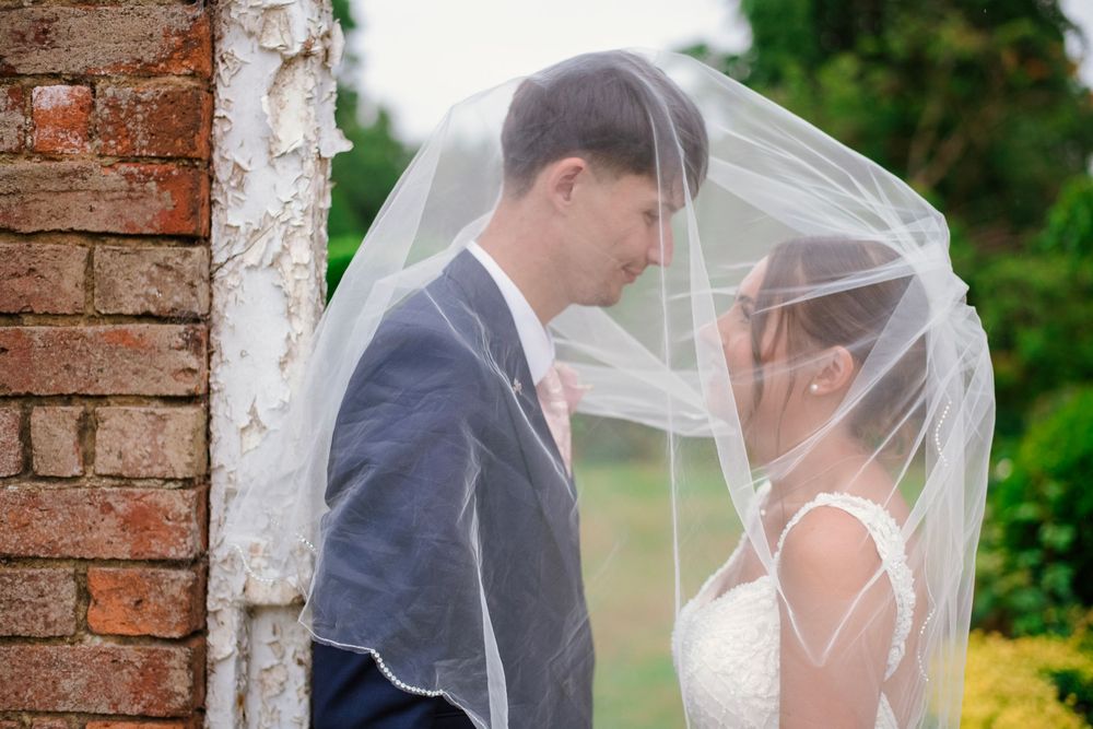 A romantic wedding couple shares an intimate moment under a flowing veil against a rustic brick wall backdrop.