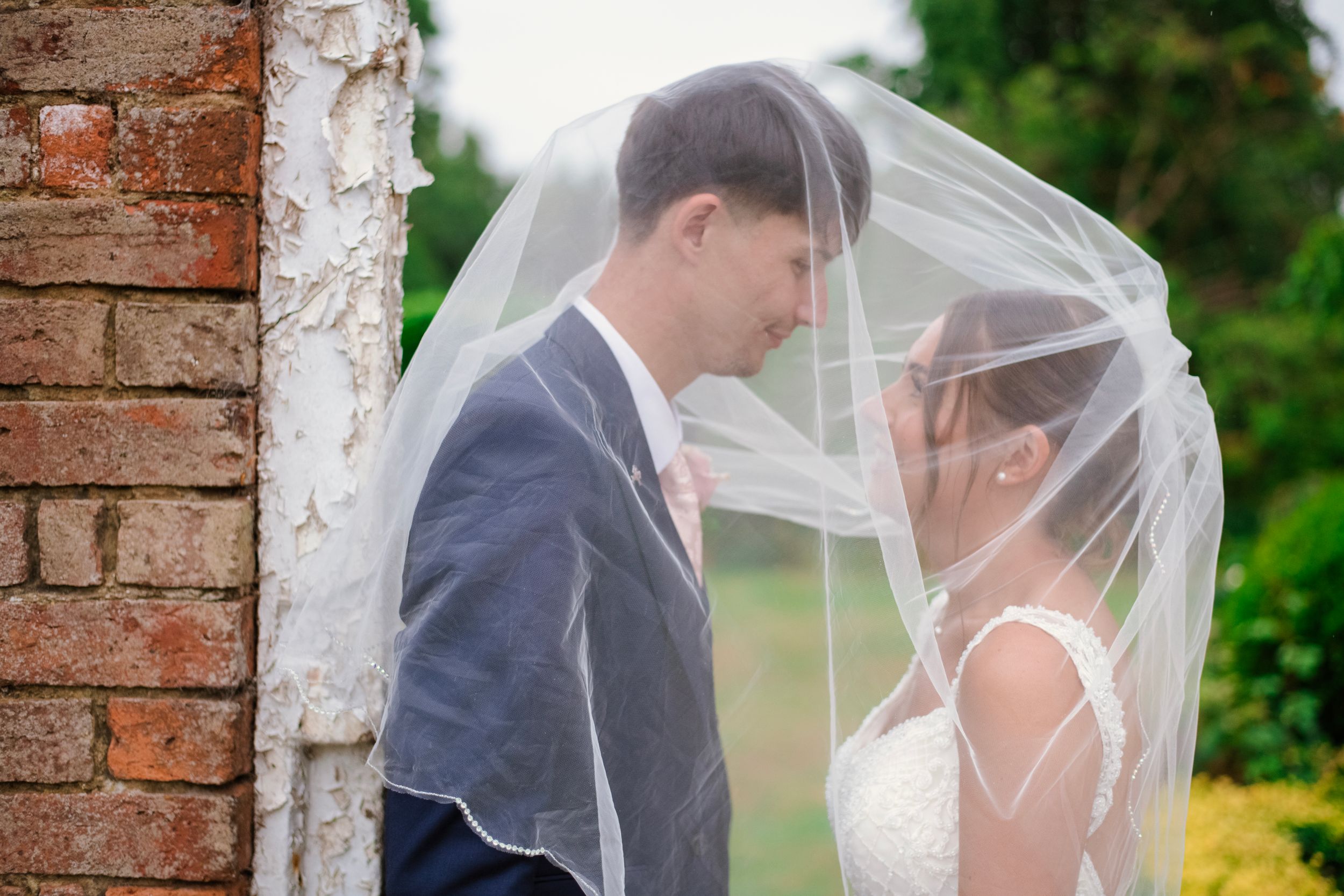 A romantic wedding couple shares an intimate moment under a flowing veil against a rustic brick wall backdrop.