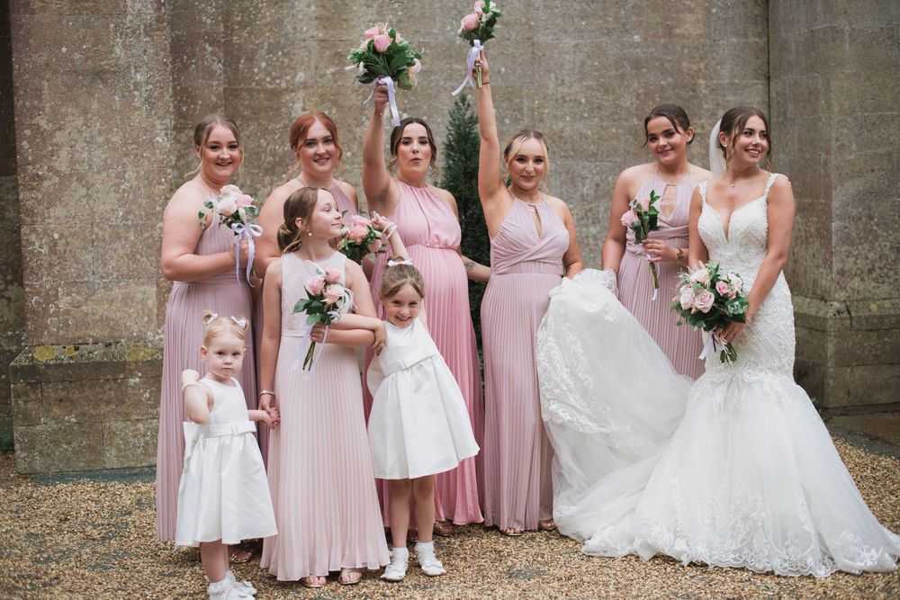 Wedding party in pink bridesmaid dresses and white gowns pose together outside stone building with bouquets.