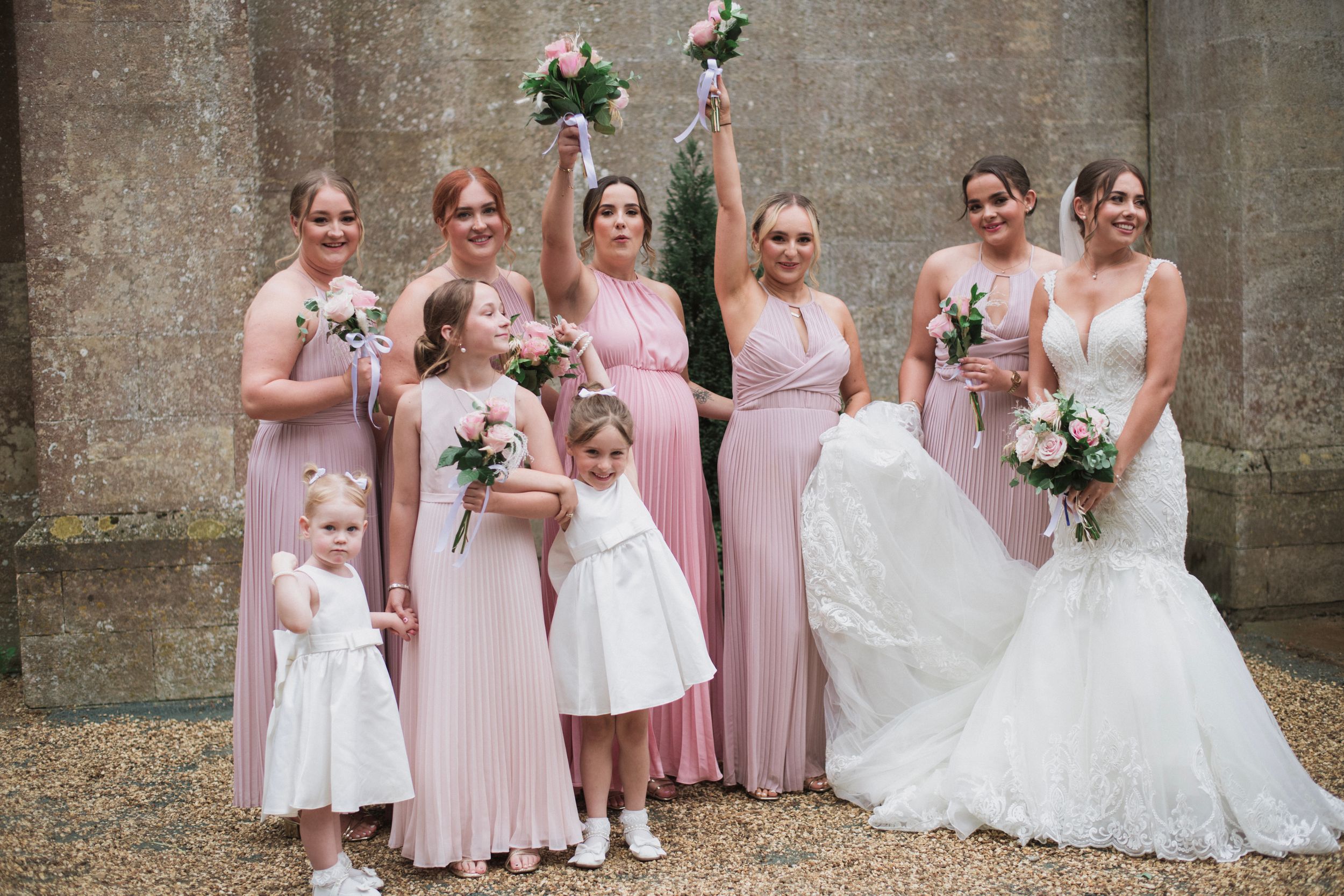 Wedding party in pink bridesmaid dresses and white gowns pose together outside stone building with bouquets.