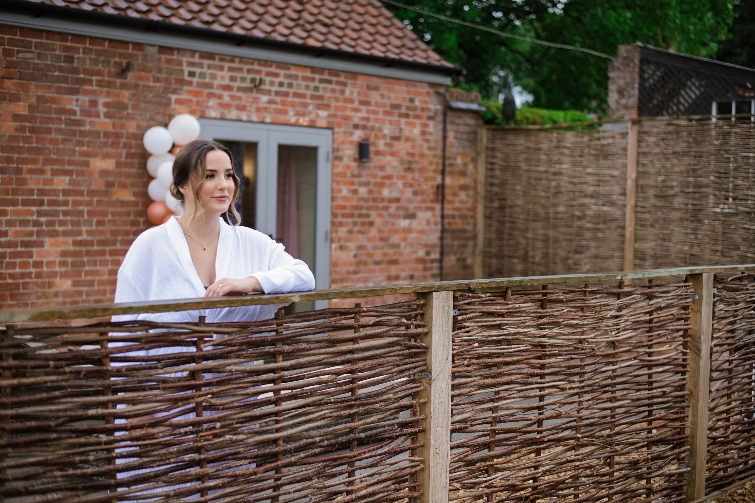 Person in white shirt leans against rustic woven fence with brick buildings and terracotta roof tiles in background.