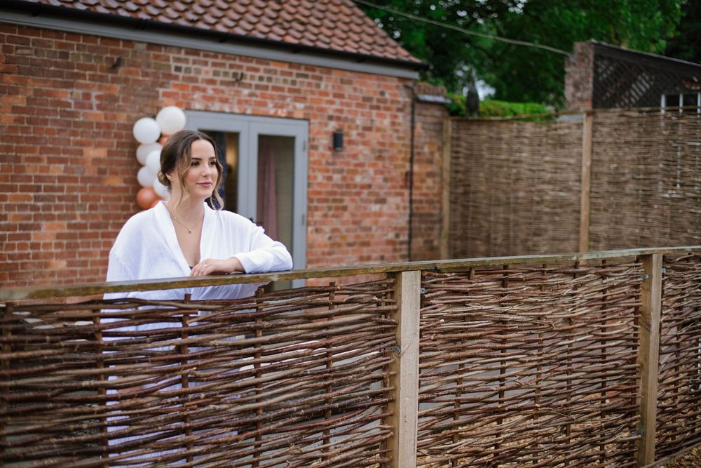 Person in white shirt leans against rustic woven fence with brick buildings and terracotta roof tiles in background.