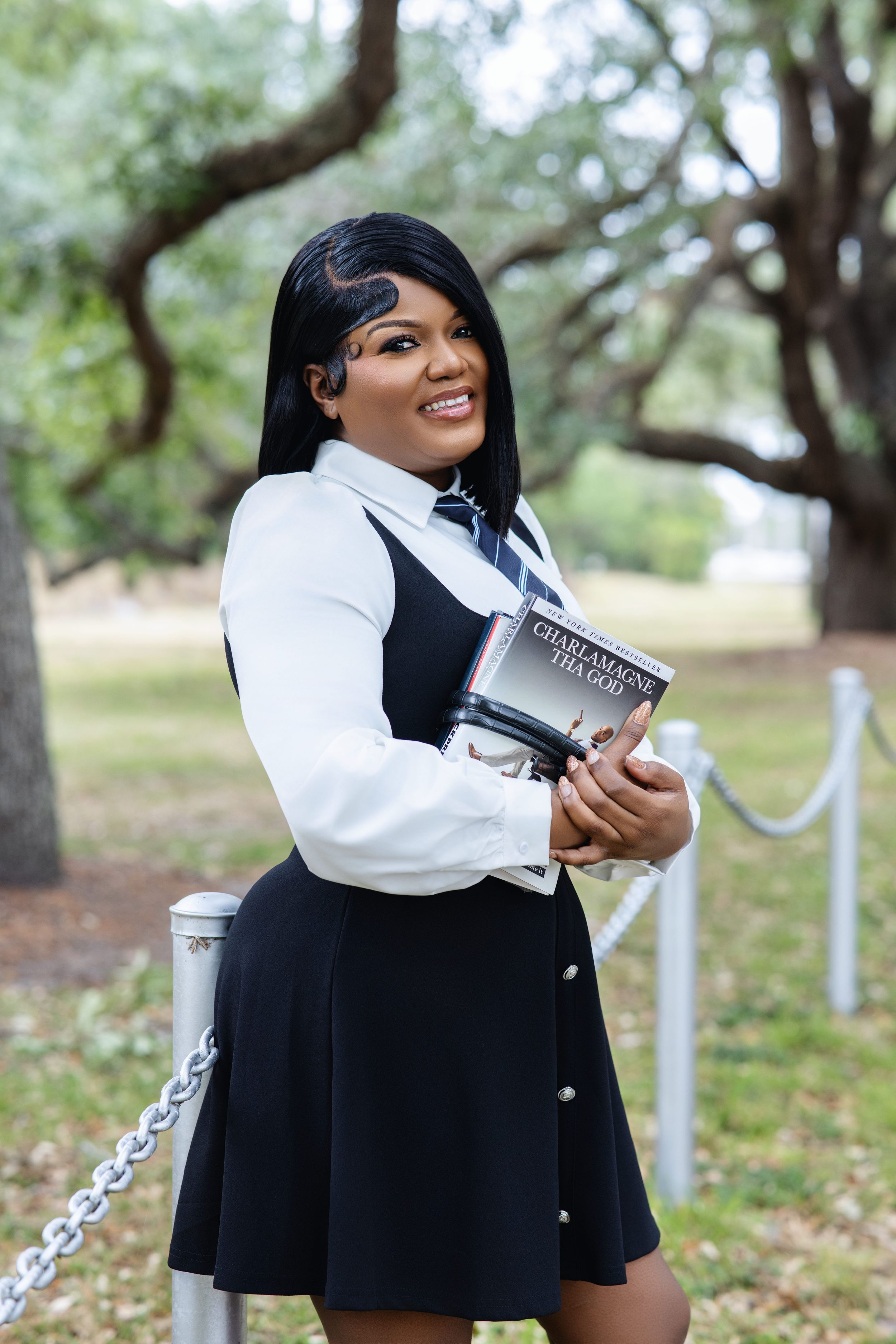 Smiling Black woman in black dress and white shirt holds Charlamagne Tha God book outdoors near chain fence.