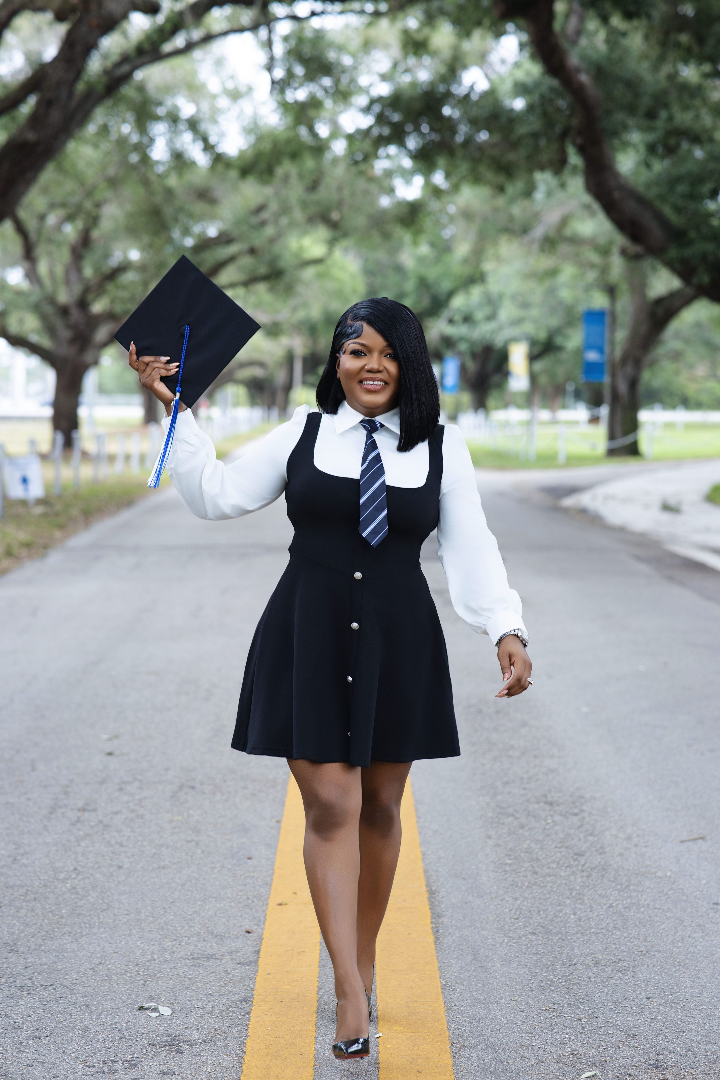 Joyful graduate holding cap while walking down tree-lined campus road.
