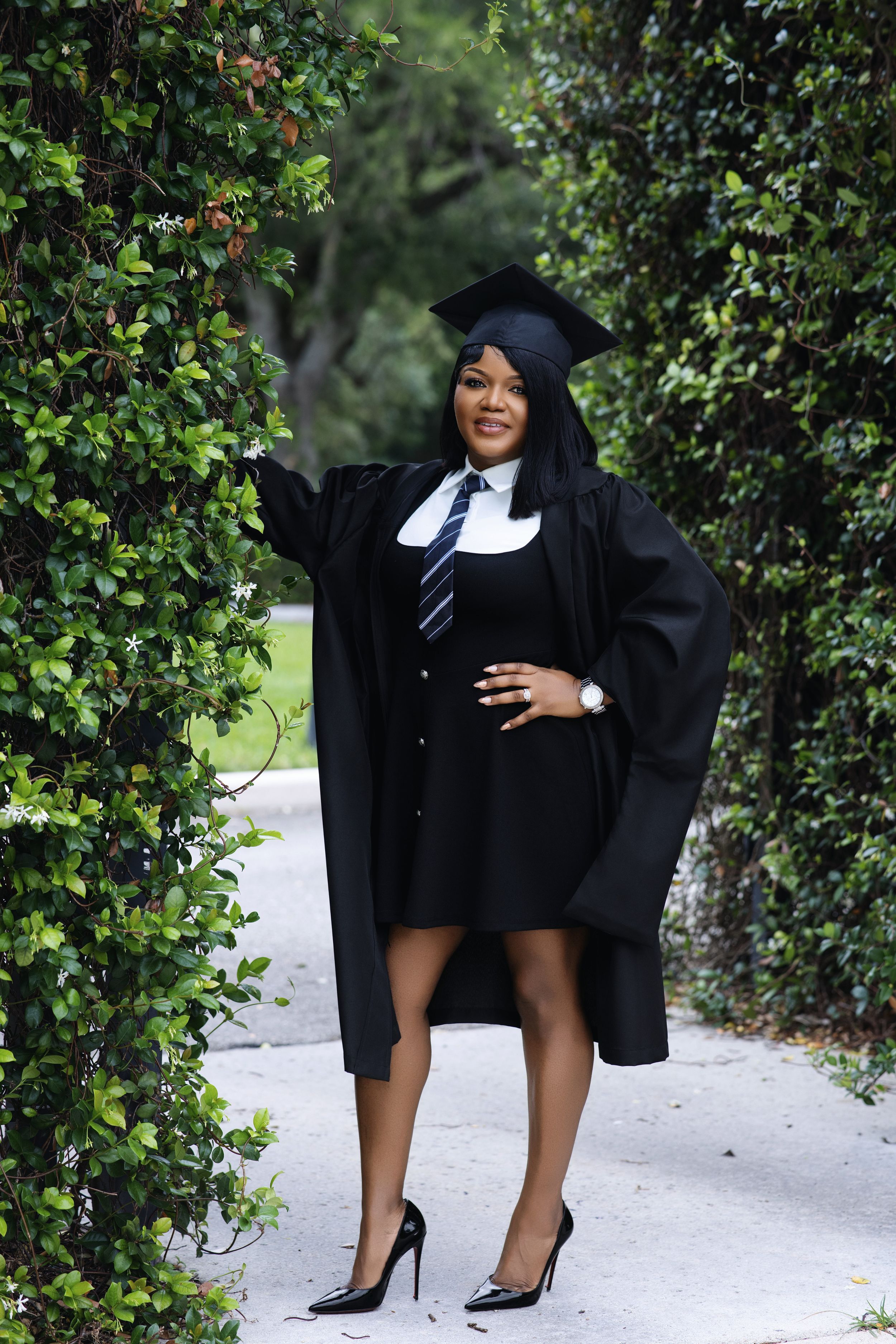 Graduate in black cap and gown posing between ivy-covered archway.