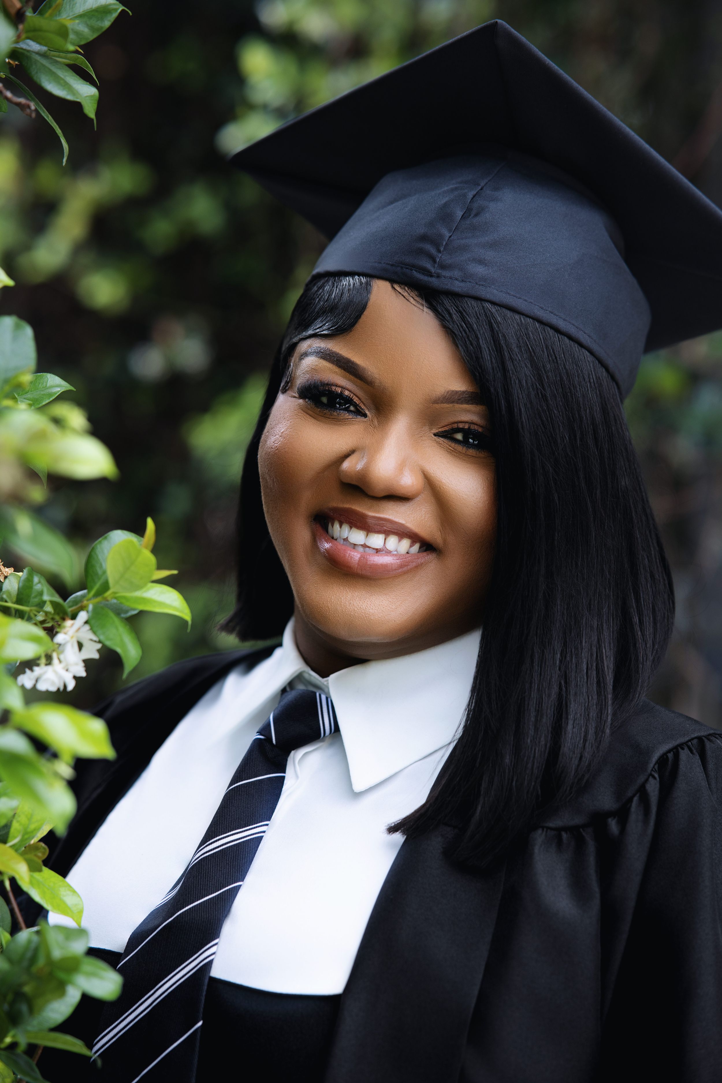 Smiling Black woman in black graduation cap and gown with white collar and striped tie poses near green foliage.