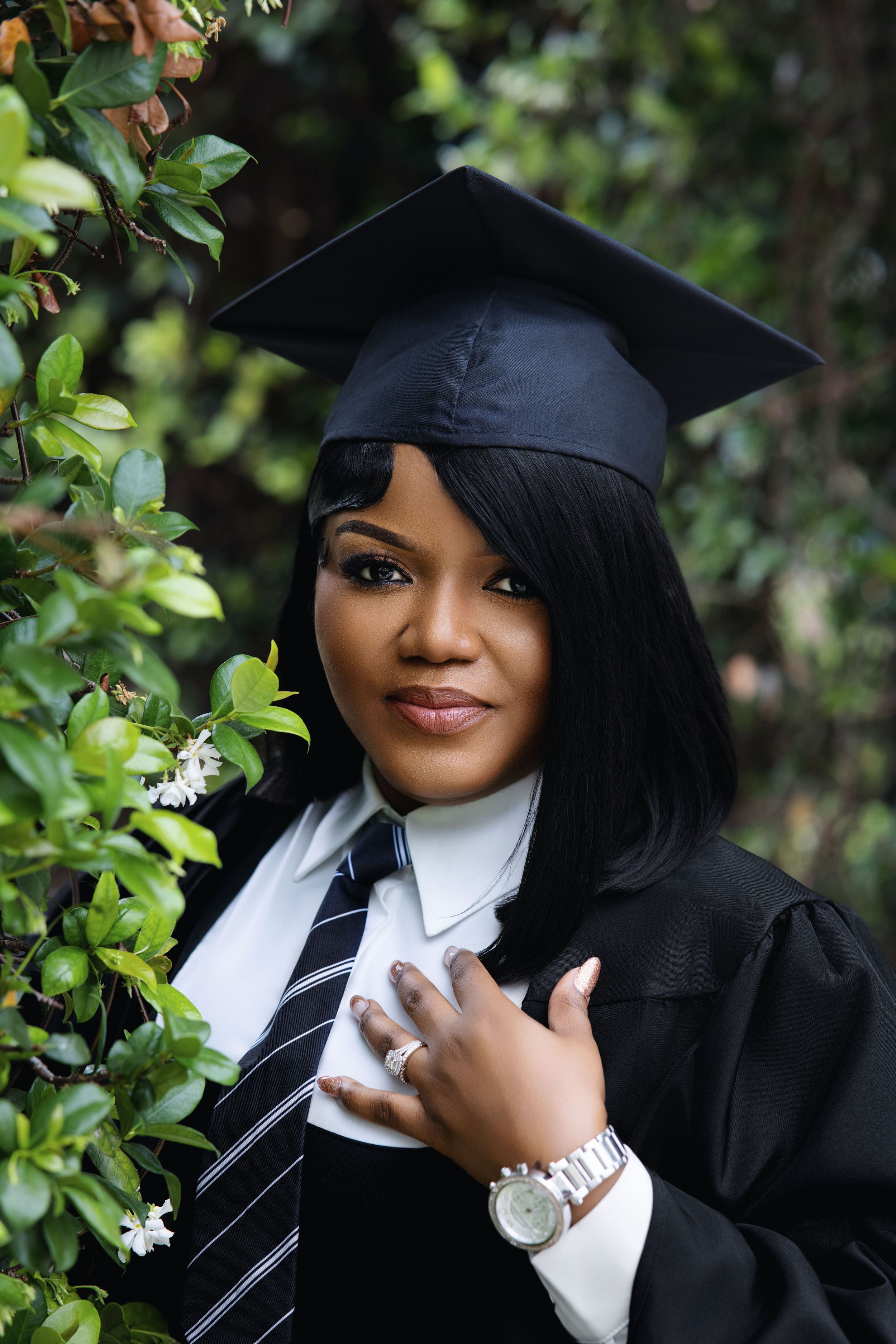 Close-up of Black woman in black graduation cap and gown with striped tie and silver watch among green leaves.