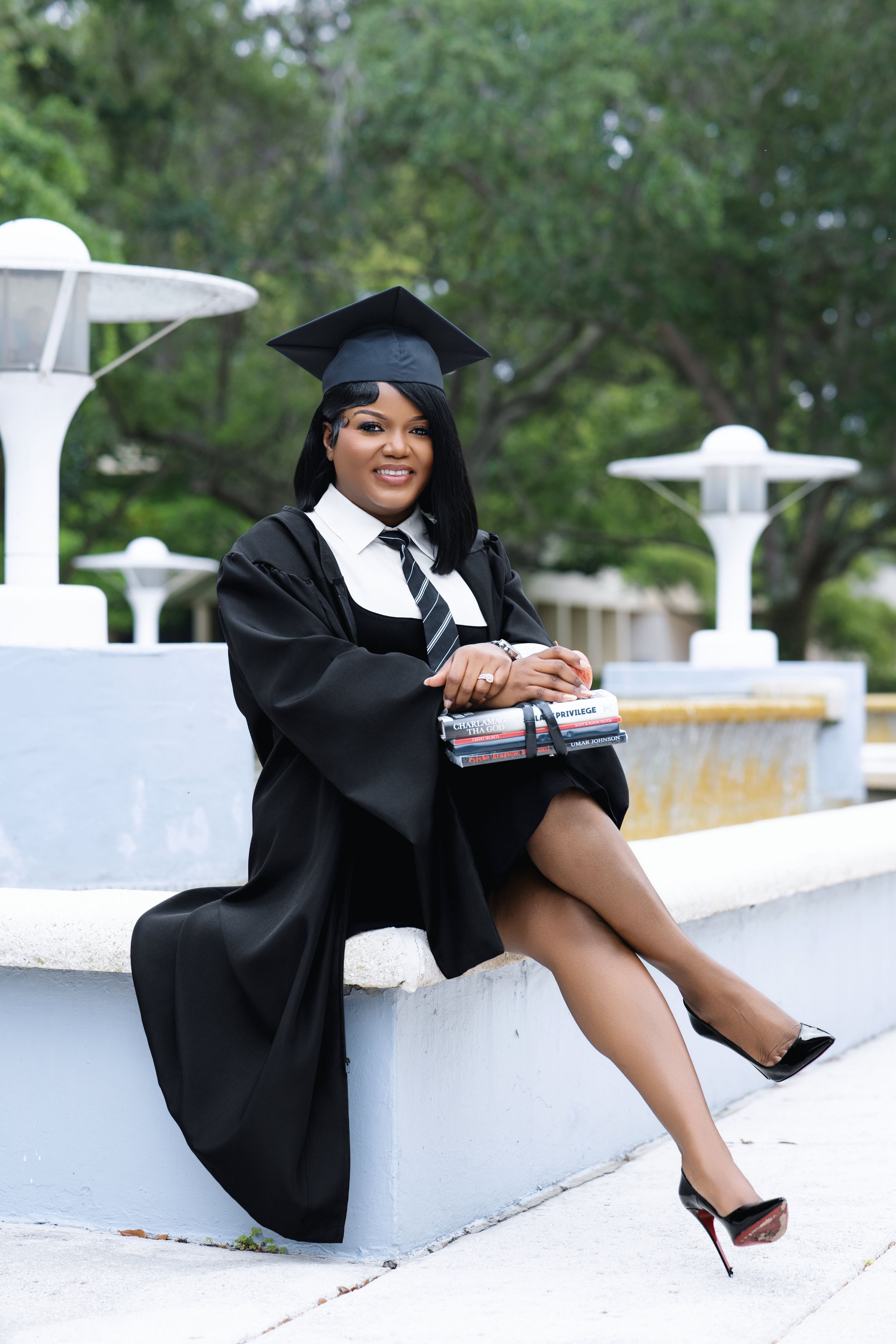 Black woman in graduation cap and gown with striped tie sits relaxed outdoors holding small wrapped gift by fountain.