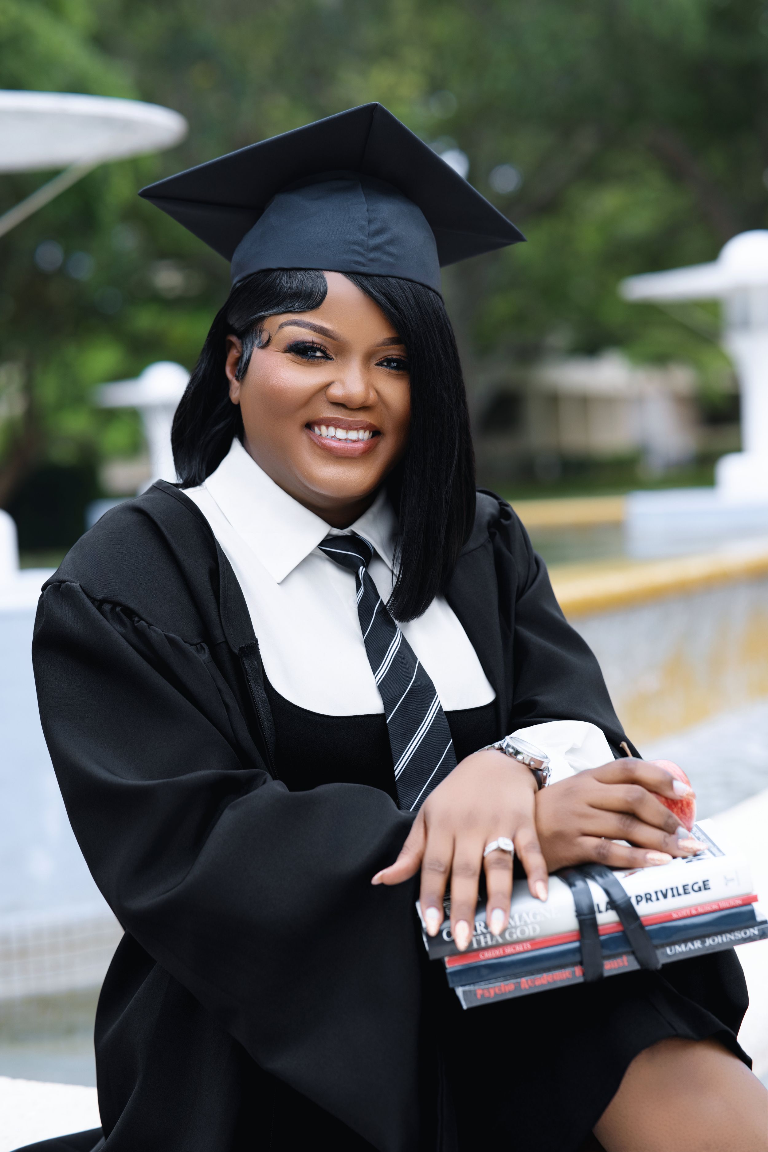Smiling graduate in black cap and gown holding books outdoors.