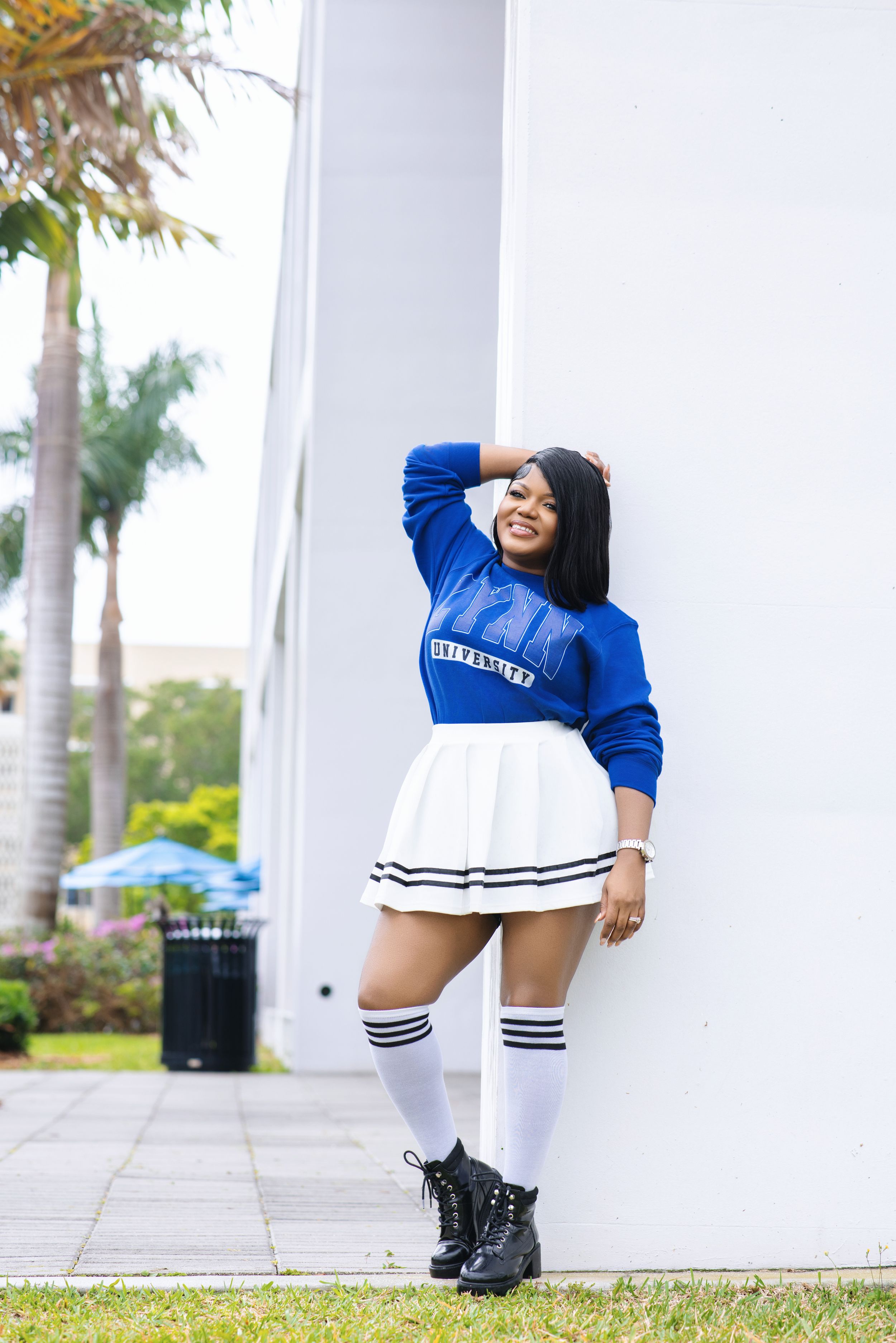 Woman in blue university top and white skirt leaning against white wall.