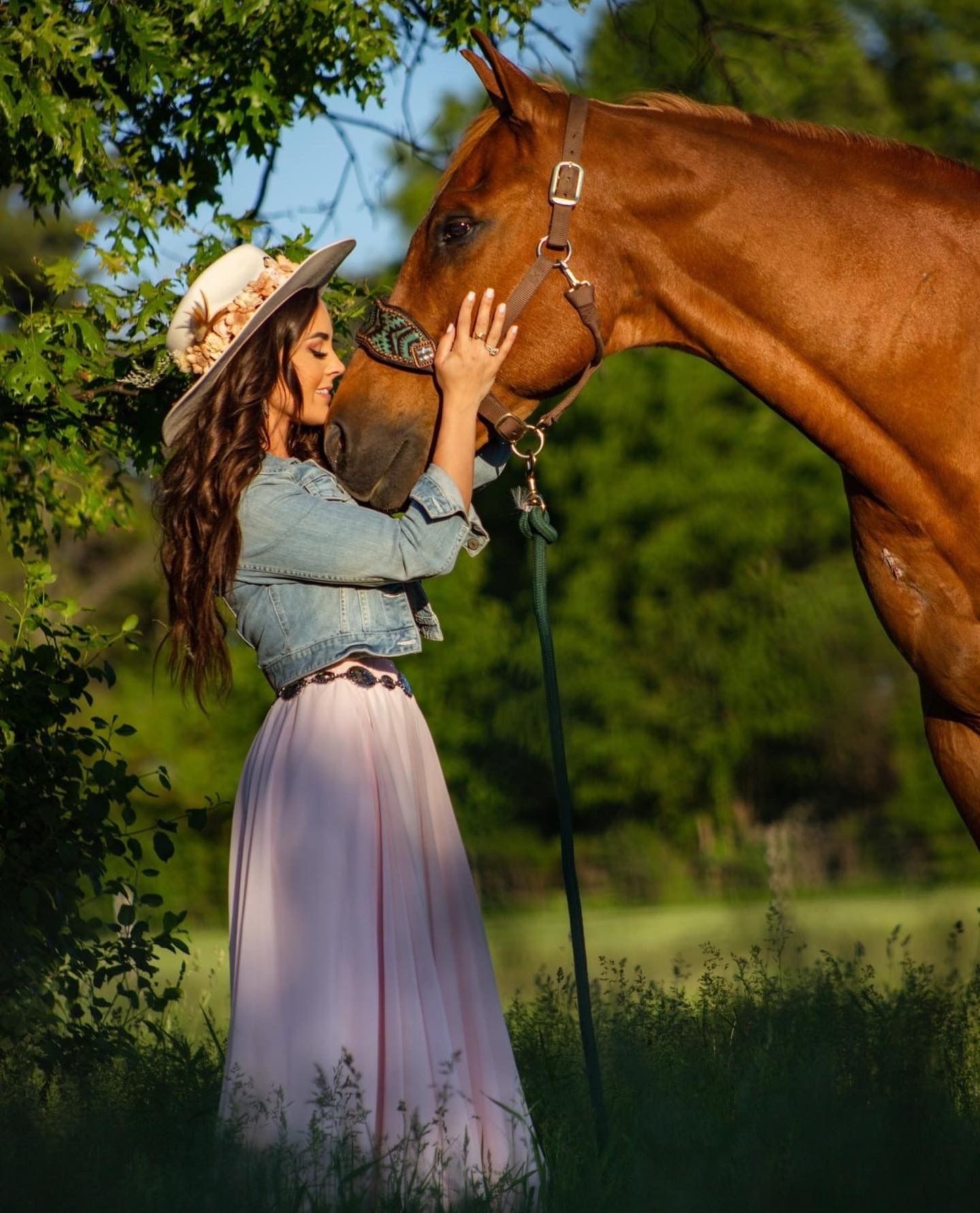 A person in a flowing pink dress stands close to a chestnut horse in a sunny outdoor field setting.