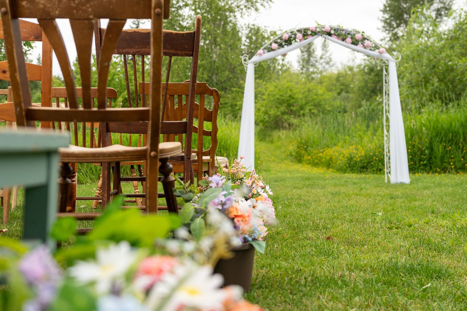 A rustic outdoor wedding ceremony setup with wooden benches and white drapes among green trees and grass. Captured by Savvy Photography